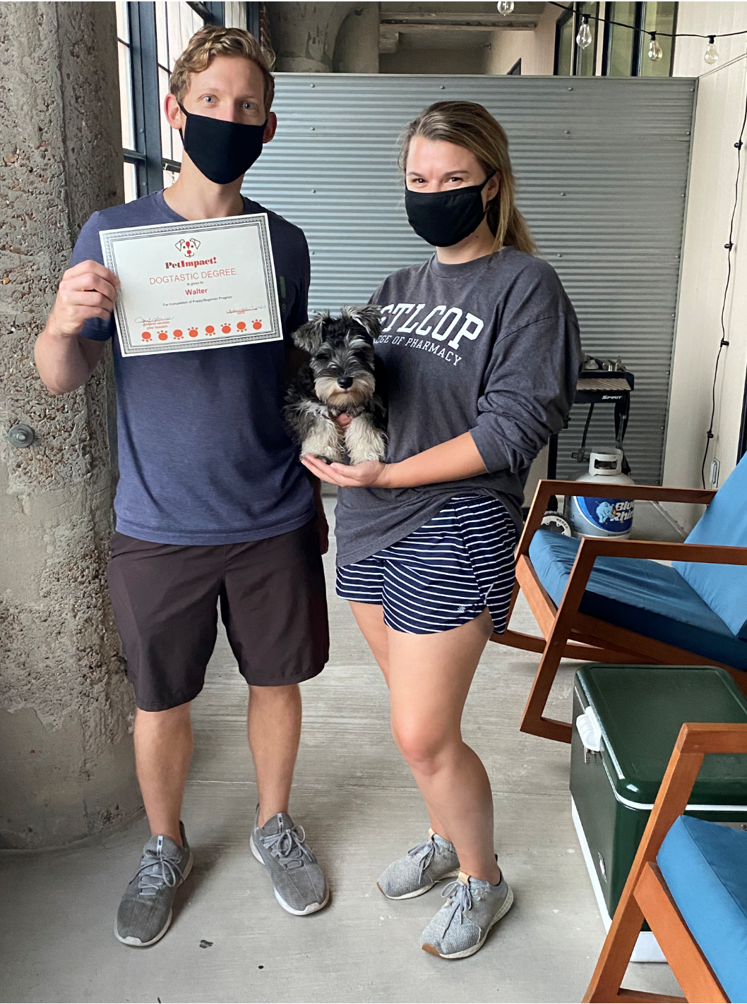 Couple holding a certificate and a small dog outside, both wearing face masks.