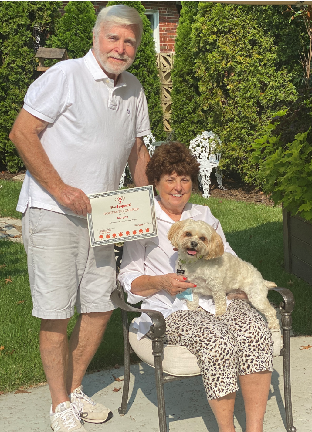 Man and woman with dog, holding a certificate outdoors. Woman seated, dog on lap, all smiling.