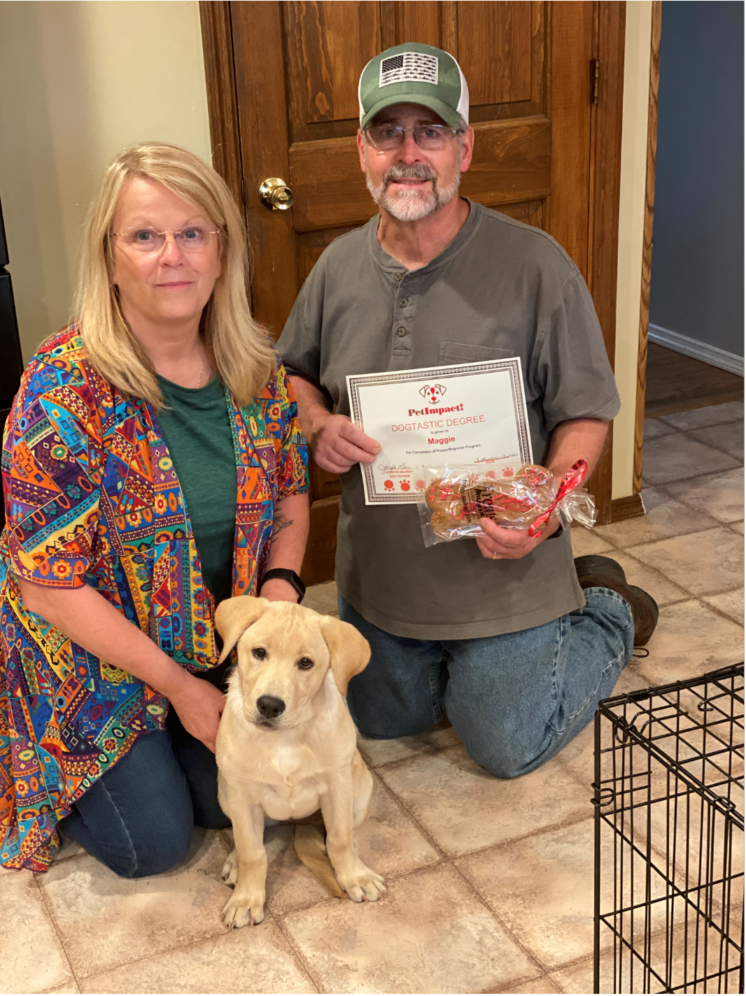 Couple with puppy, holding certificate and treats, kneeling on tile floor near a crate.