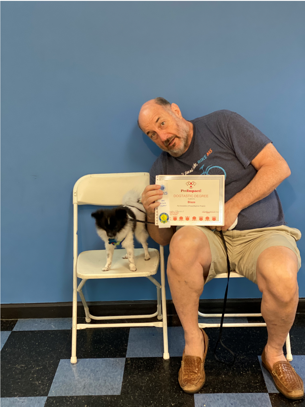 Man seated, holding certificate next to small dog on chair; blue wall background.
