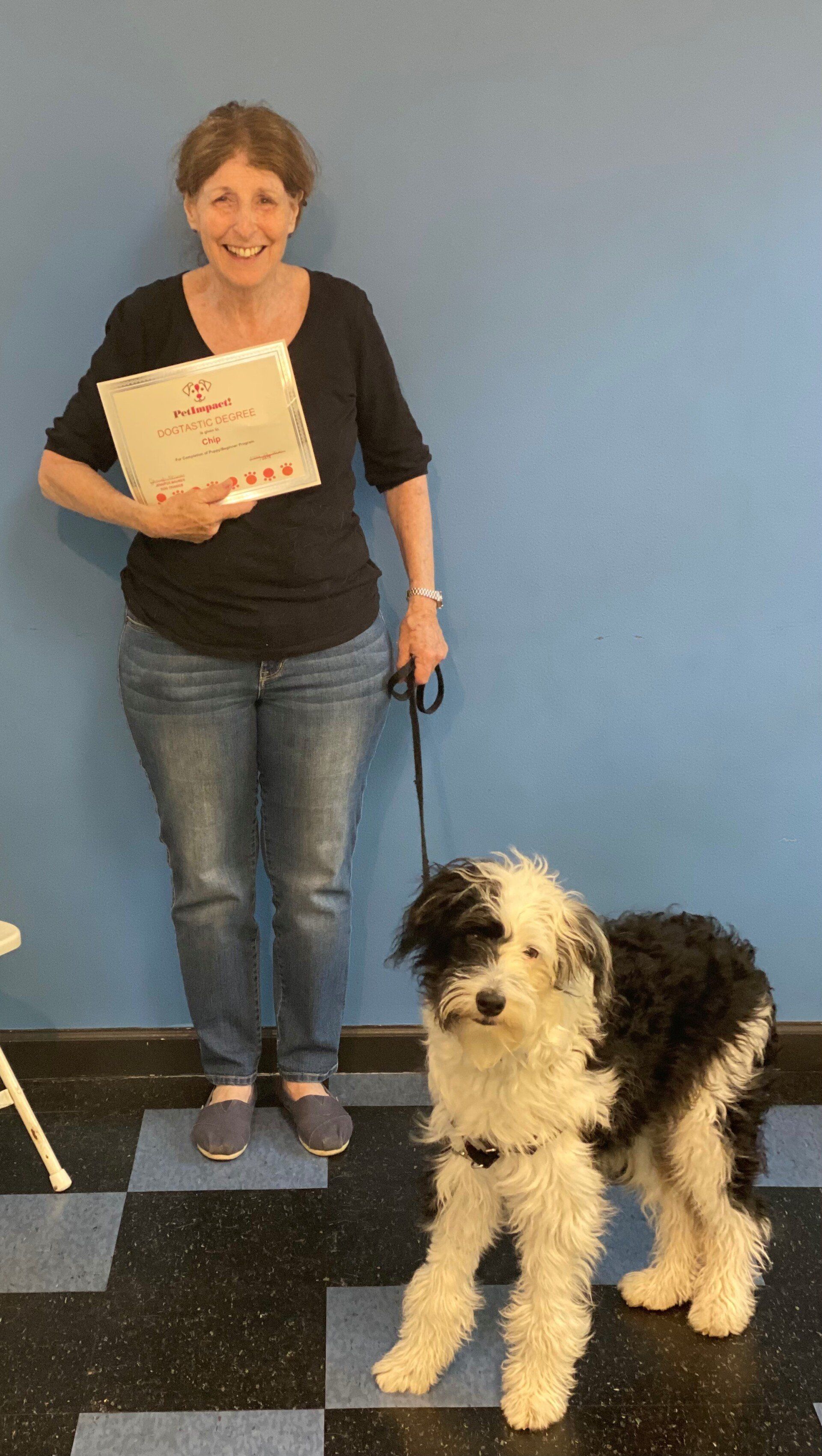 Woman with a dog holds a certificate in front of a blue wall; dog is black and white, standing on a checkered floor.