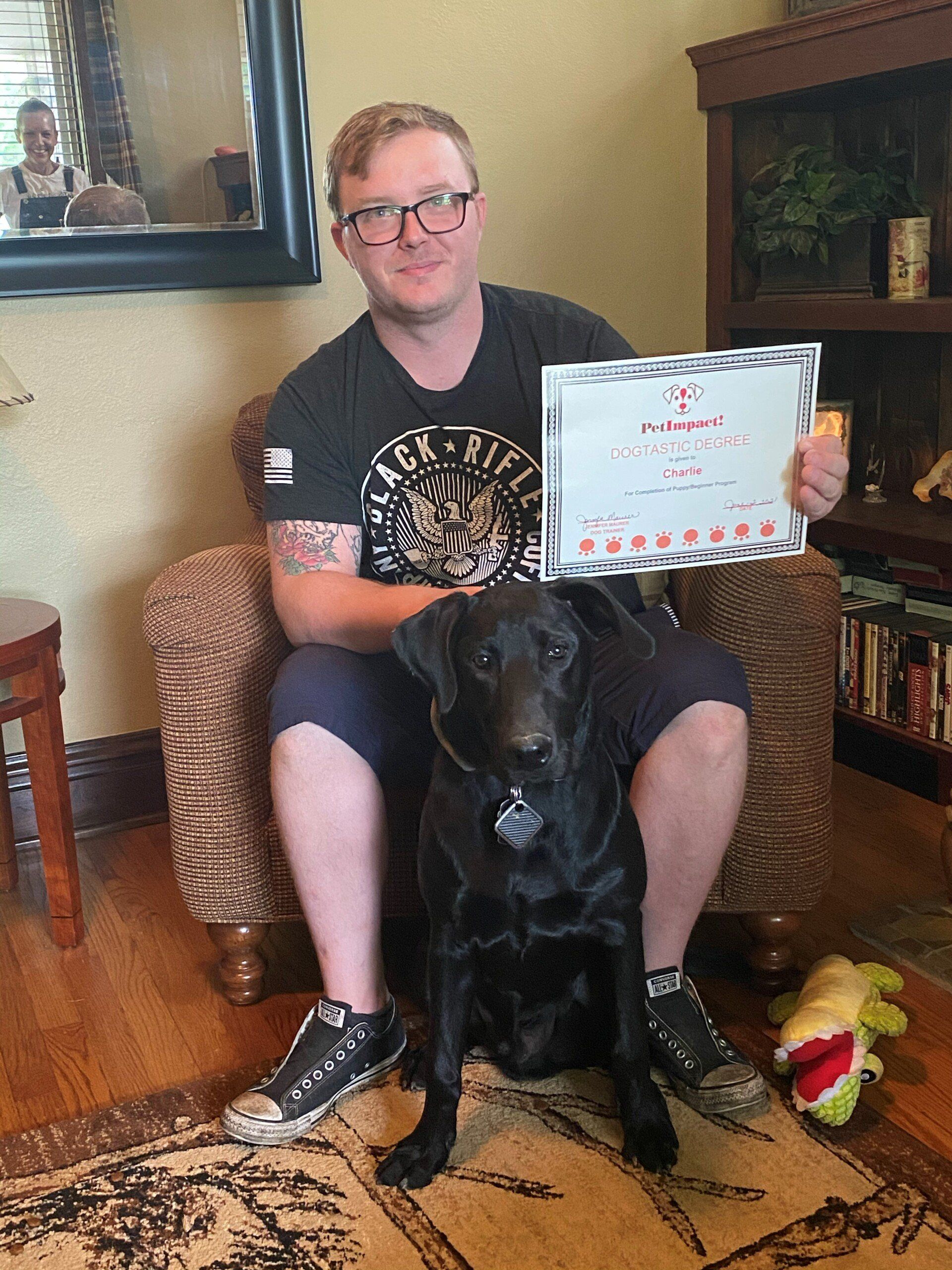 Man with glasses and black dog seated, holding framed paper; living room setting.