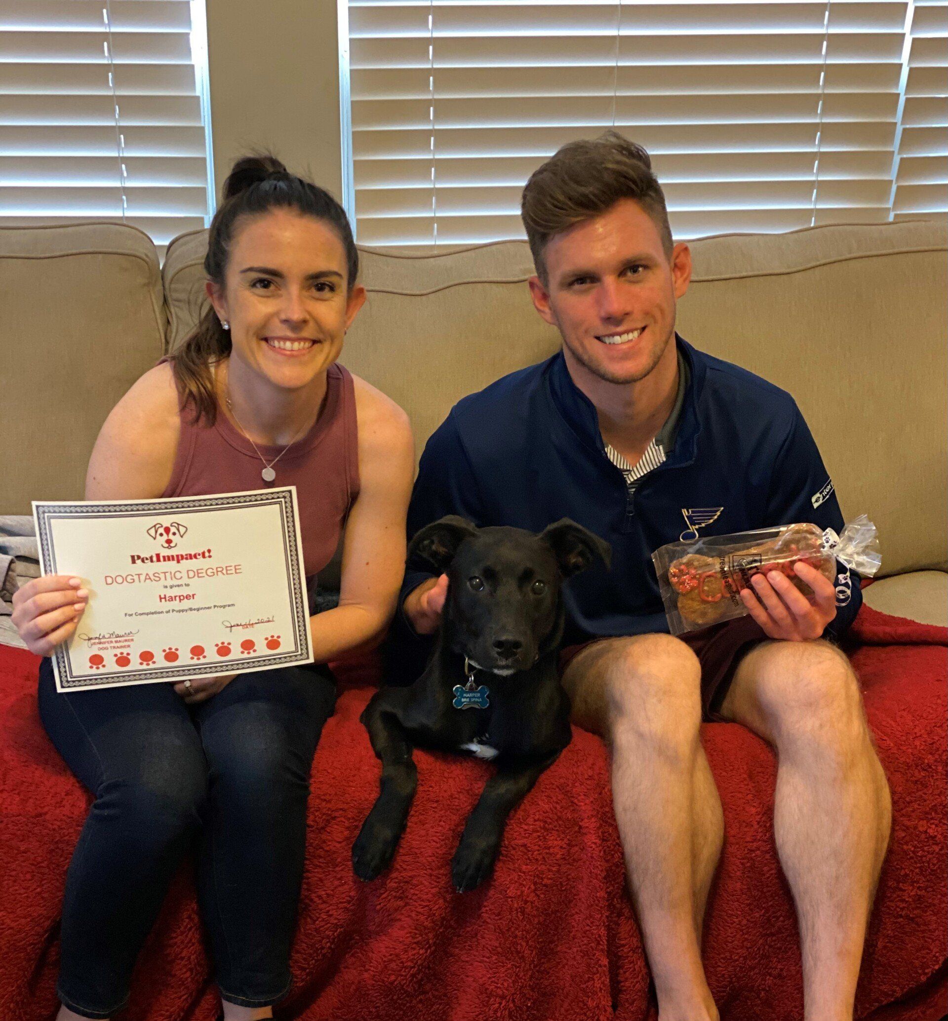 Couple with dog holding a certificate and food, sitting on a red blanket. They are smiling.