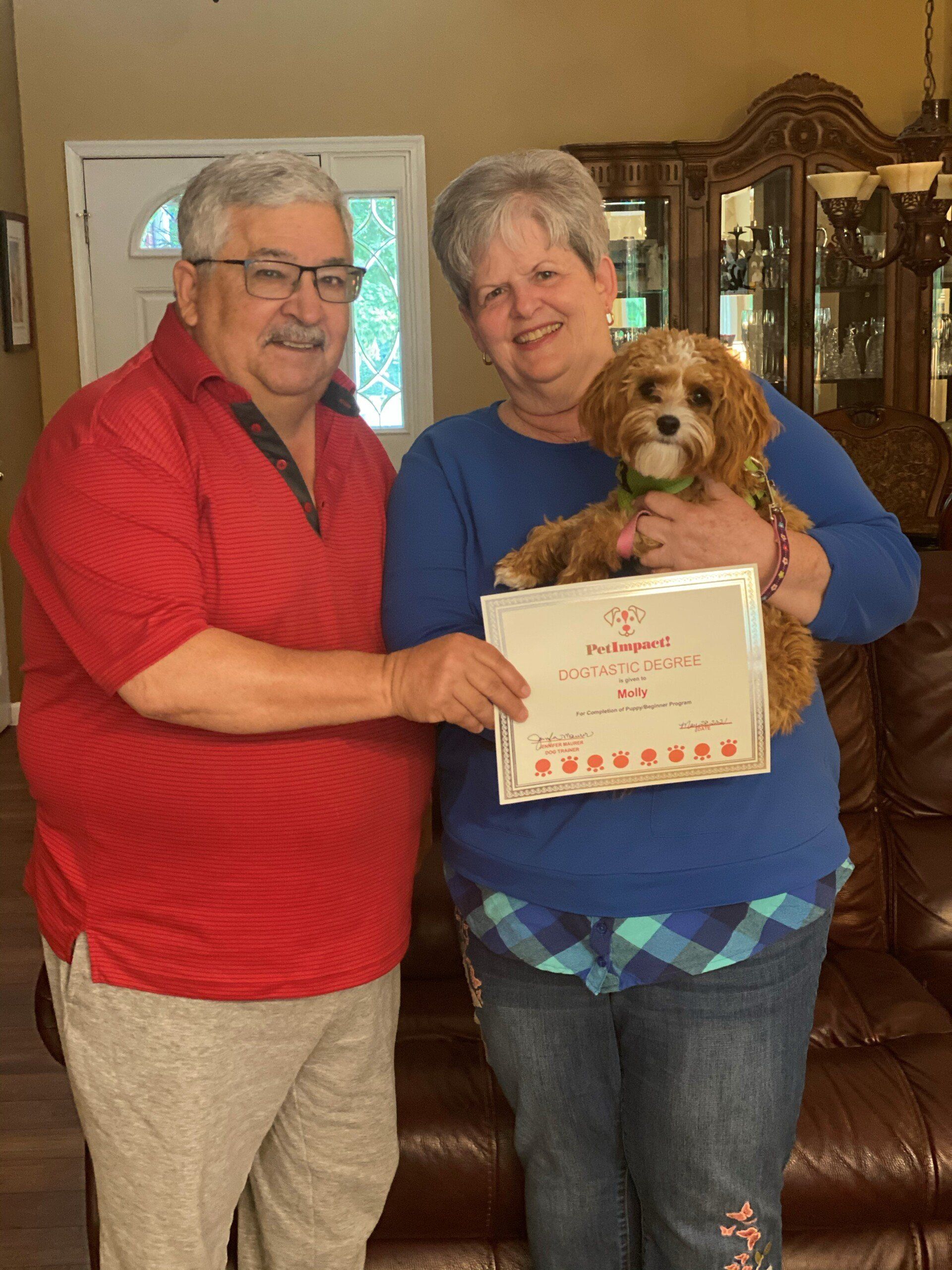 Couple holding a puppy and certificate inside a home. The man wears red, the woman blue.