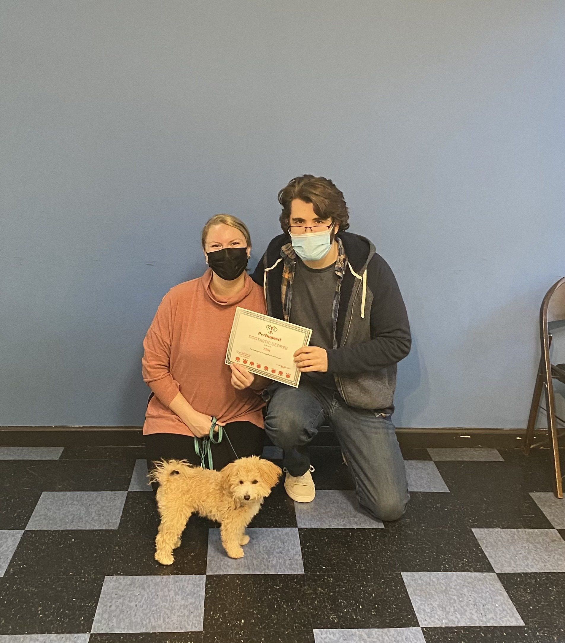 People with dog, holding certificate, by blue wall, checkered floor. They wear masks.