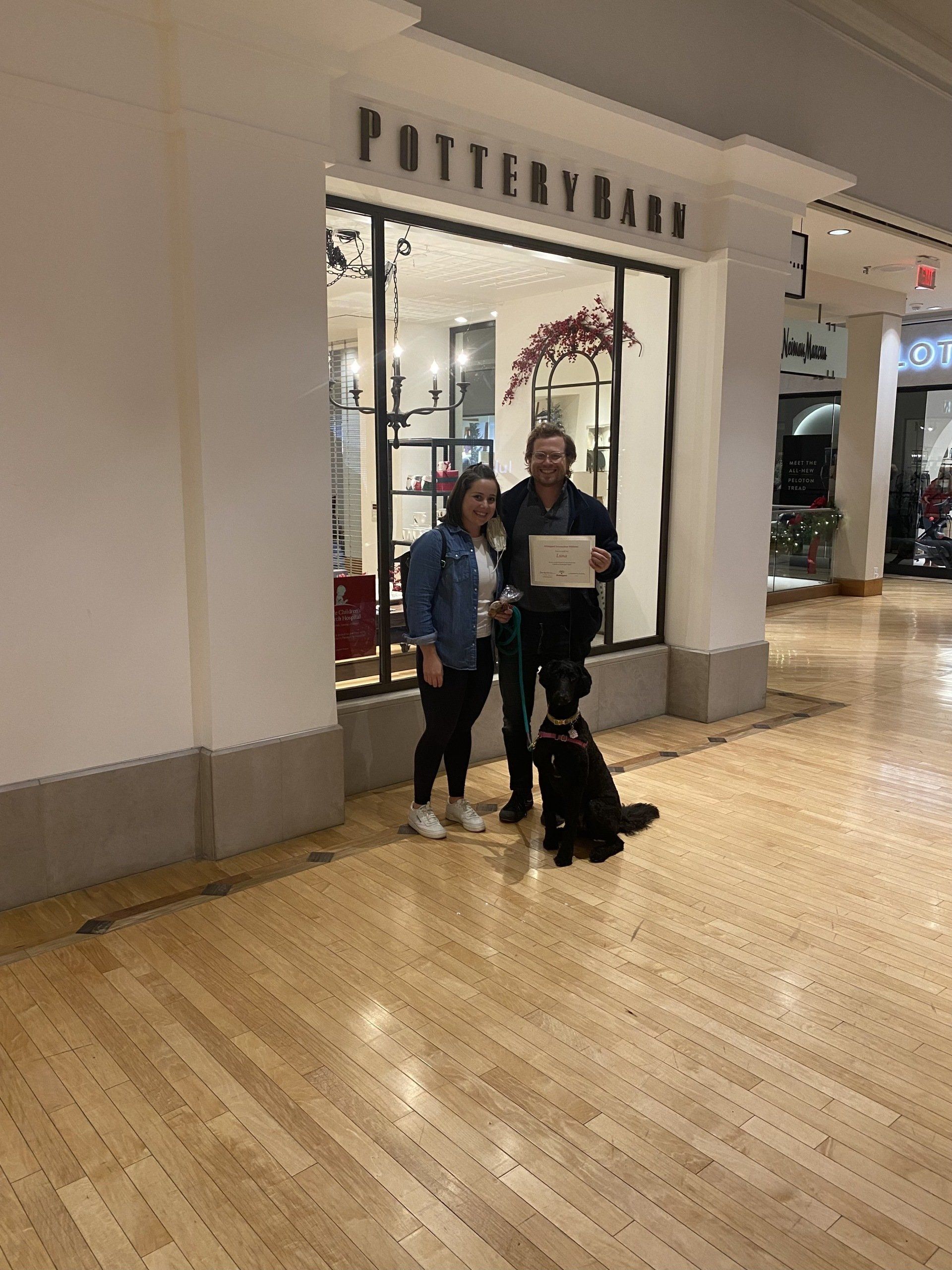 Couple and black dog pose in front of Pottery Barn store in a mall. Man holds a document.