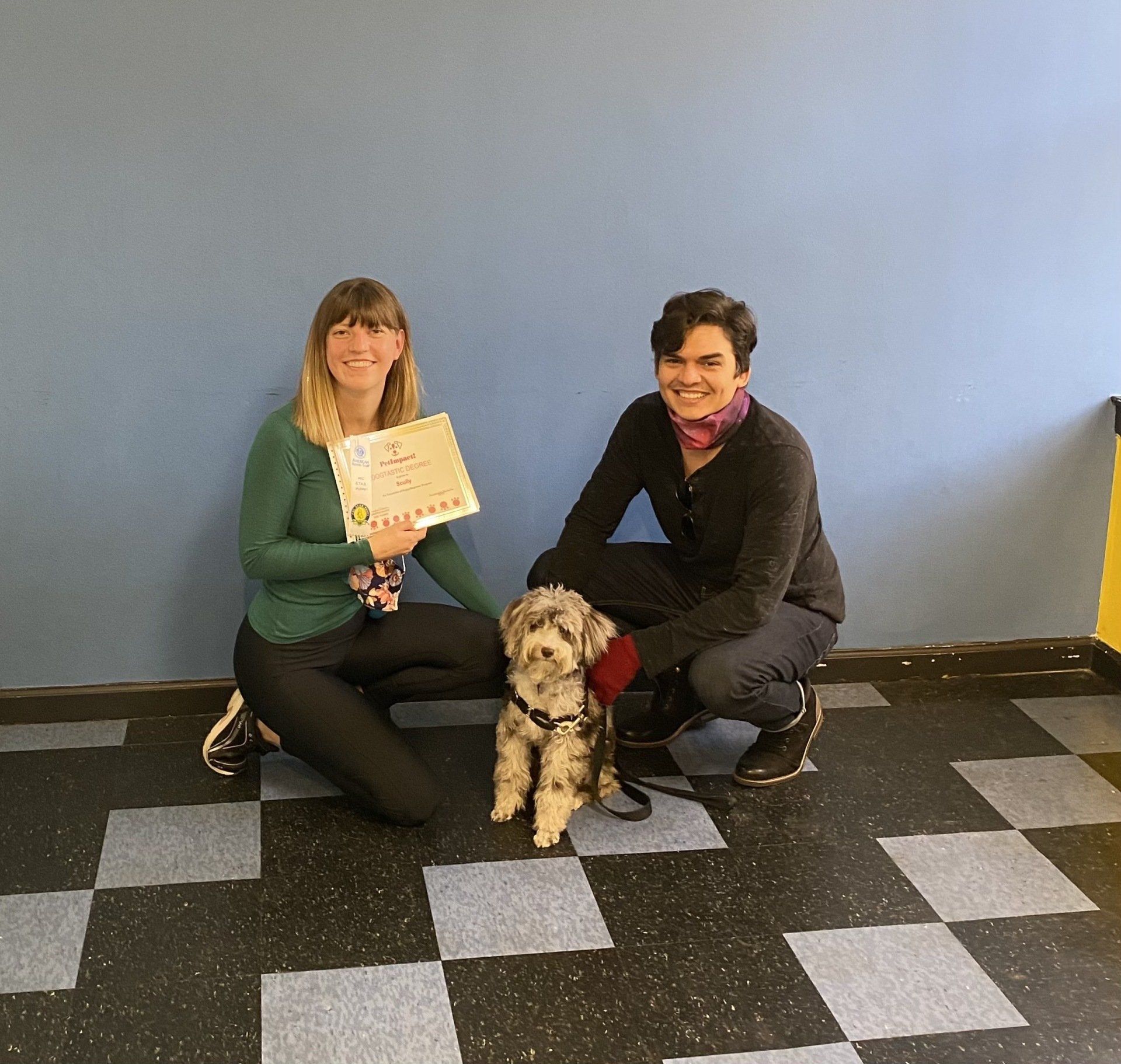 Two people crouch with a small dog. One holds a certificate. Blue wall, black and gray floor.