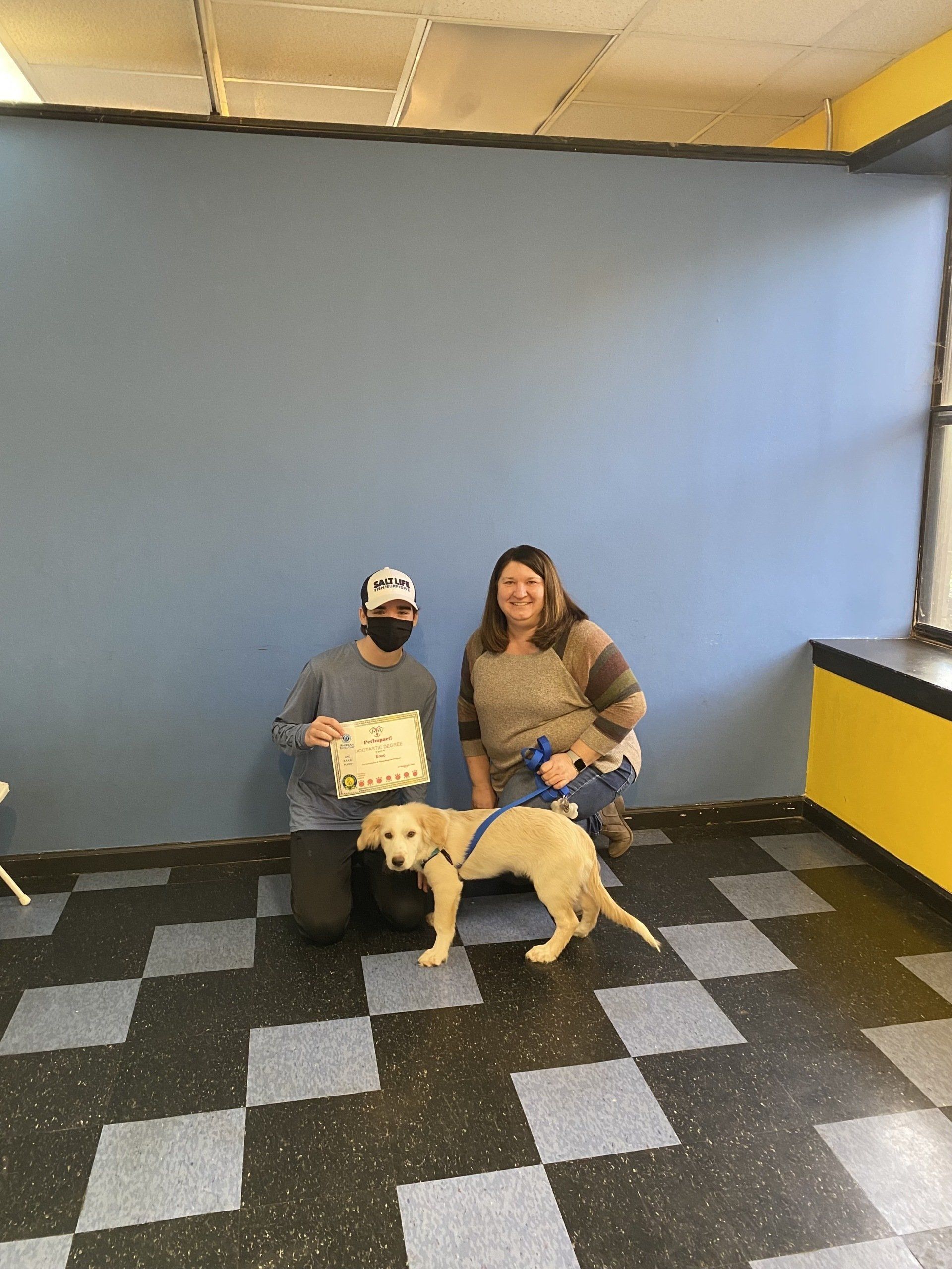 Person kneeling with a puppy and another person, posing next to a blue wall. Black and gray checkered floor.