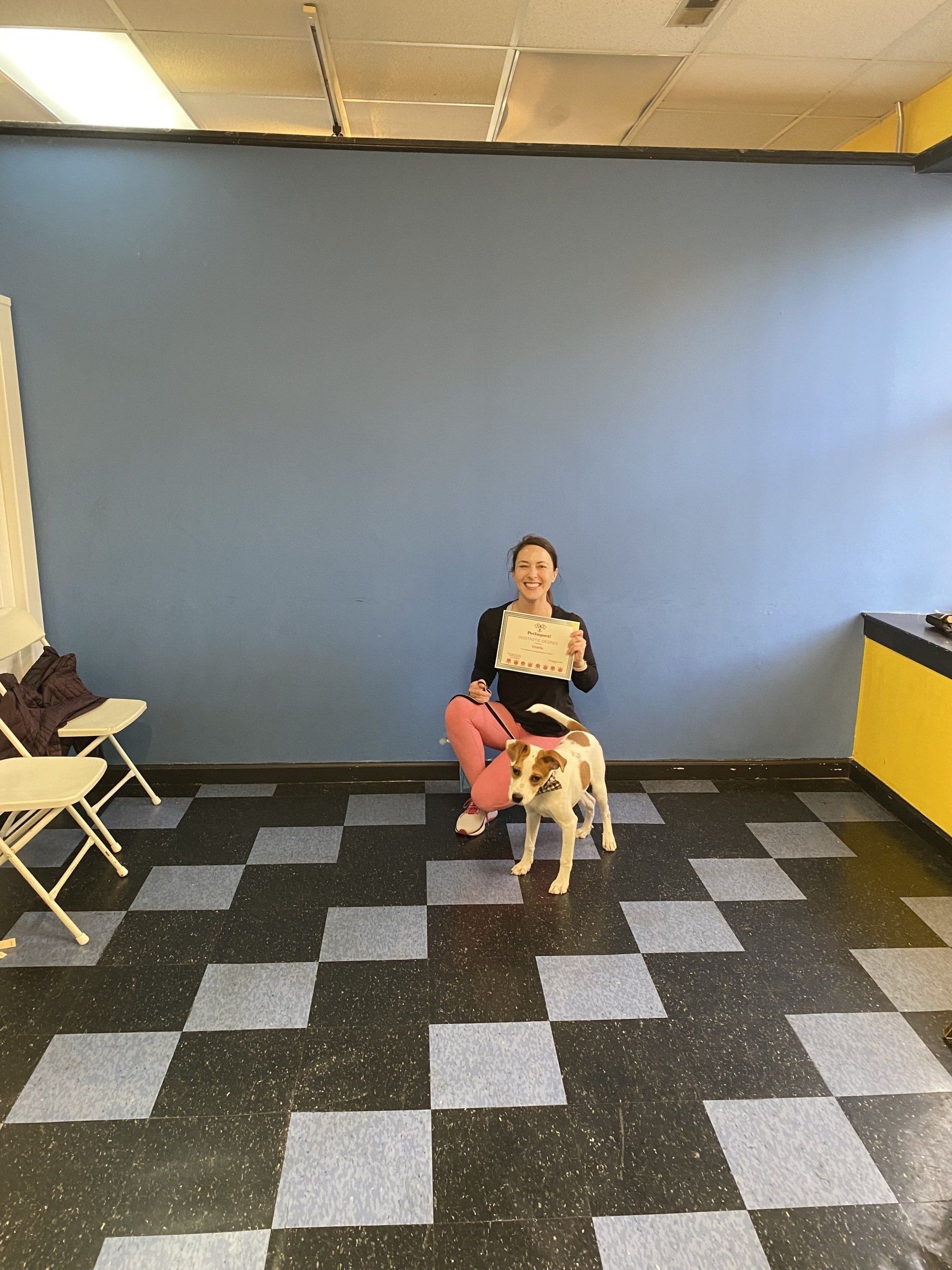 Woman seated, holding paper, poses with dog on black and blue checkered floor, blue wall in background.