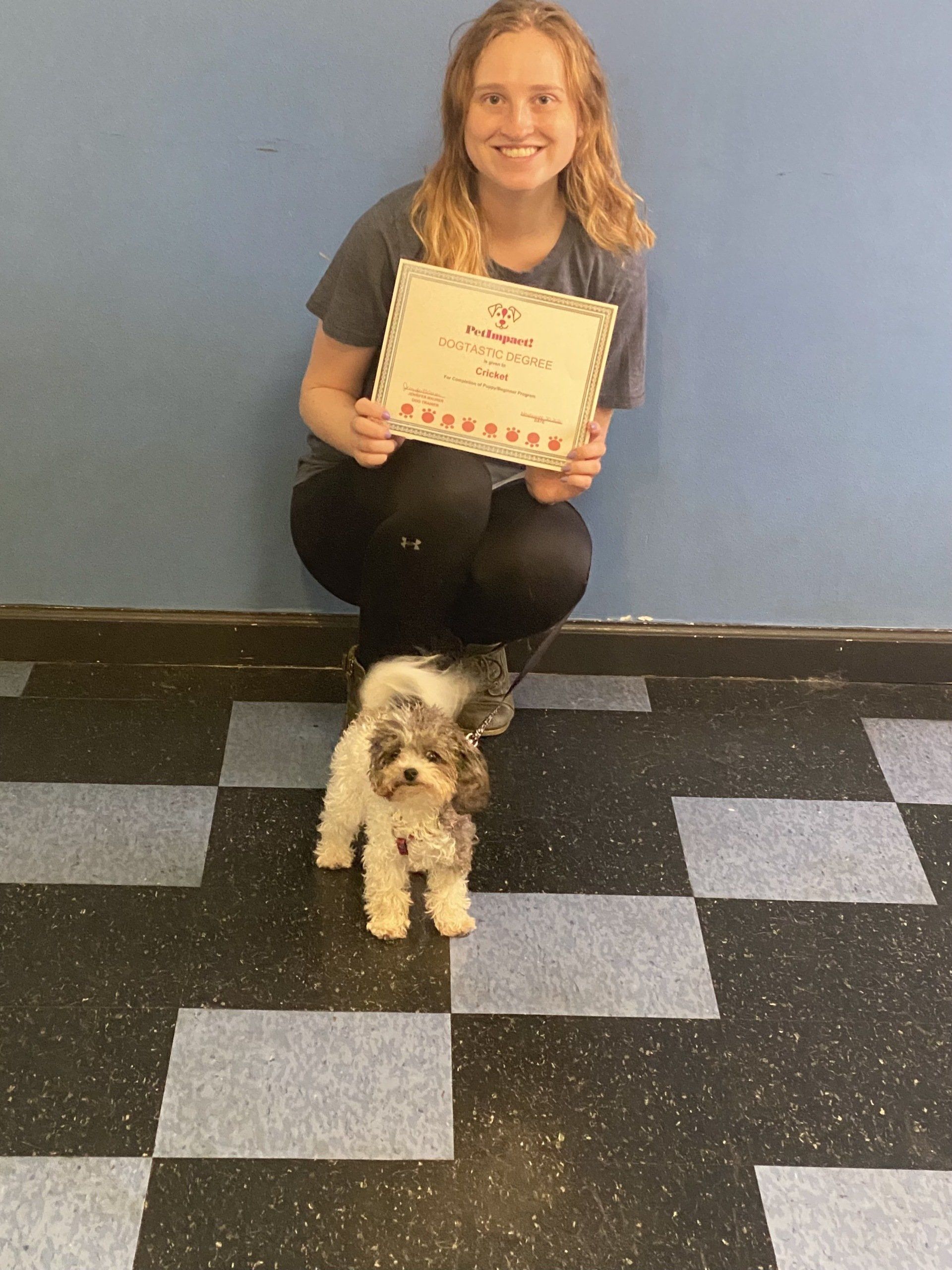 Woman with dog holding certificate; blue wall backdrop, checkered floor.