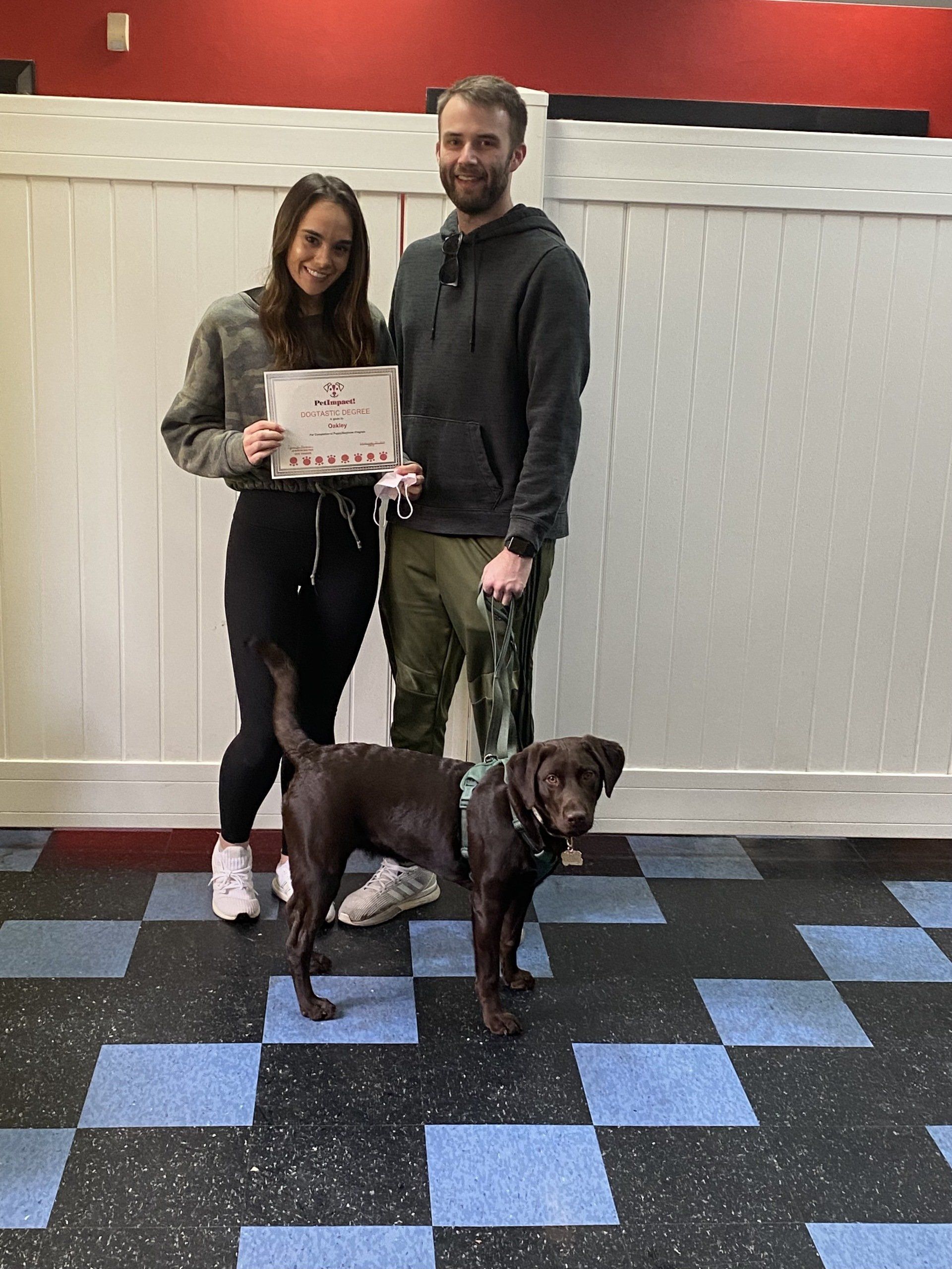 A couple and a brown dog pose together, holding a certificate. They stand in front of a white fence, on a blue-tiled floor.
