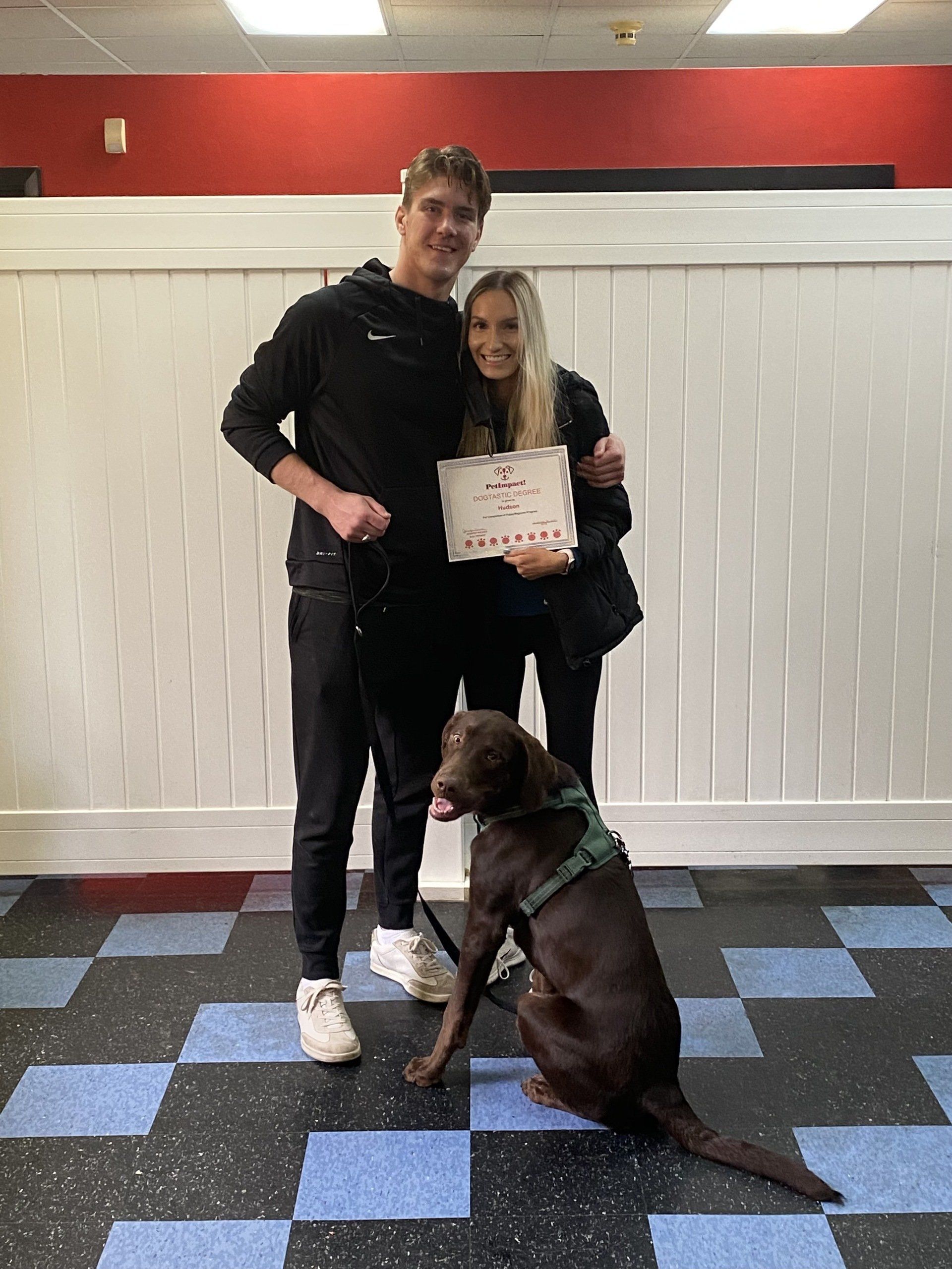 A couple and a brown dog posing, holding a sign. The couple is standing with the dog on a leash.