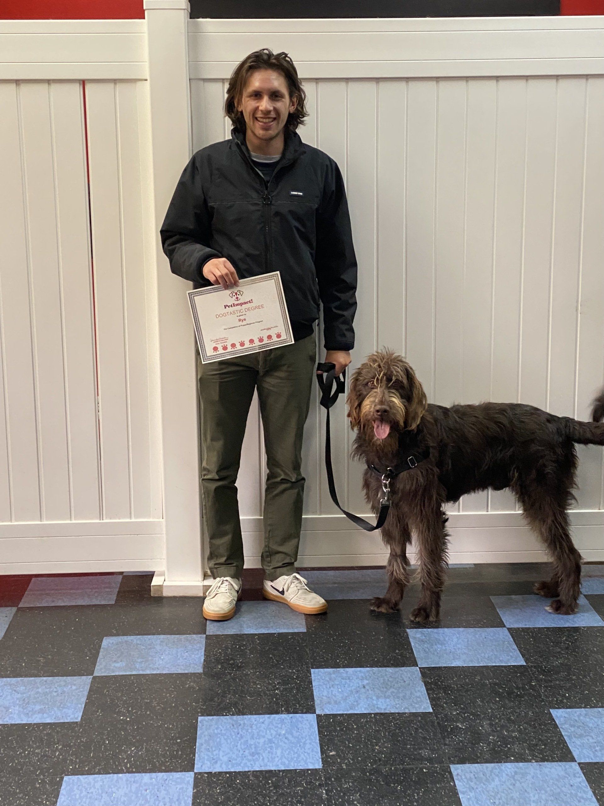 Man and brown dog stand on checkered floor, holding a sign. The man wears a black jacket, green pants, and white shoes.