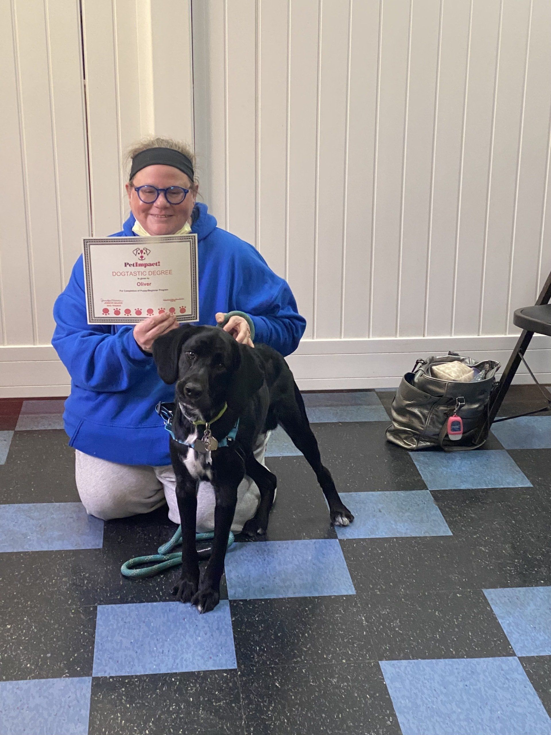 Woman kneeling with a black dog, holding a certificate. They are on a blue and gray tiled floor.