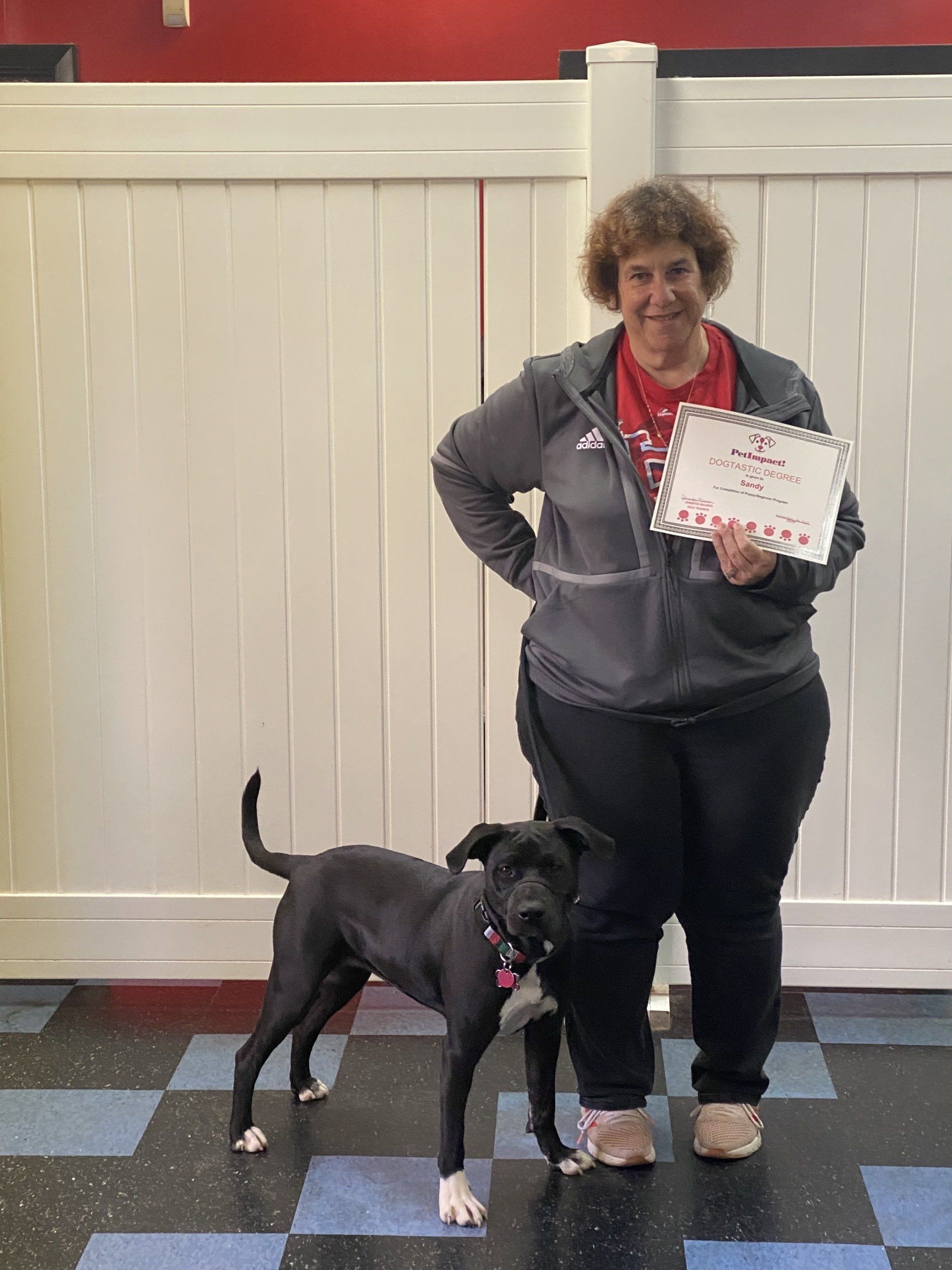 Woman in gray jacket stands next to a black dog. They are in front of a white wall.