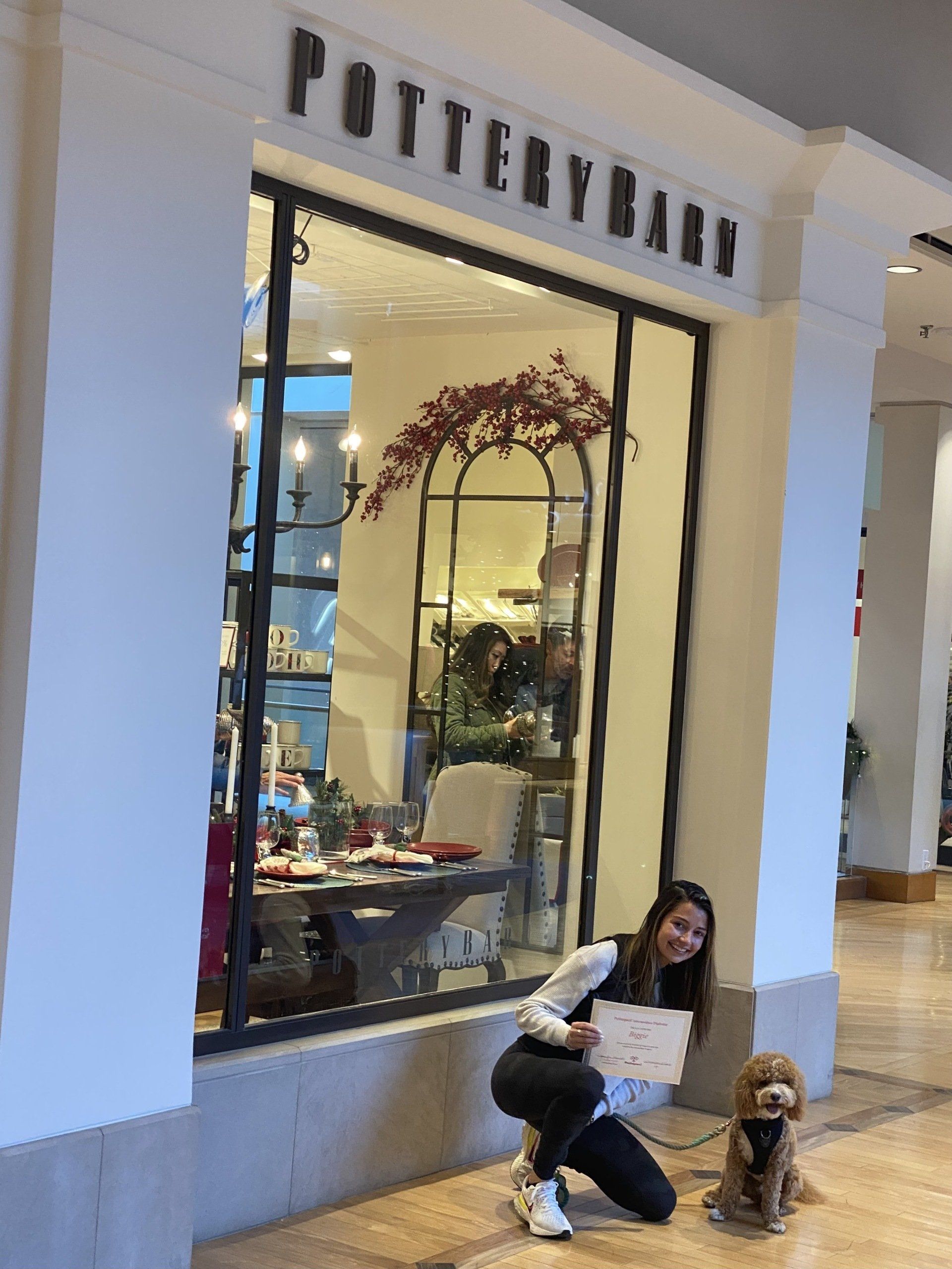 Woman and small dog pose outside Pottery Barn store in a mall. The woman holds a sign, smiling.