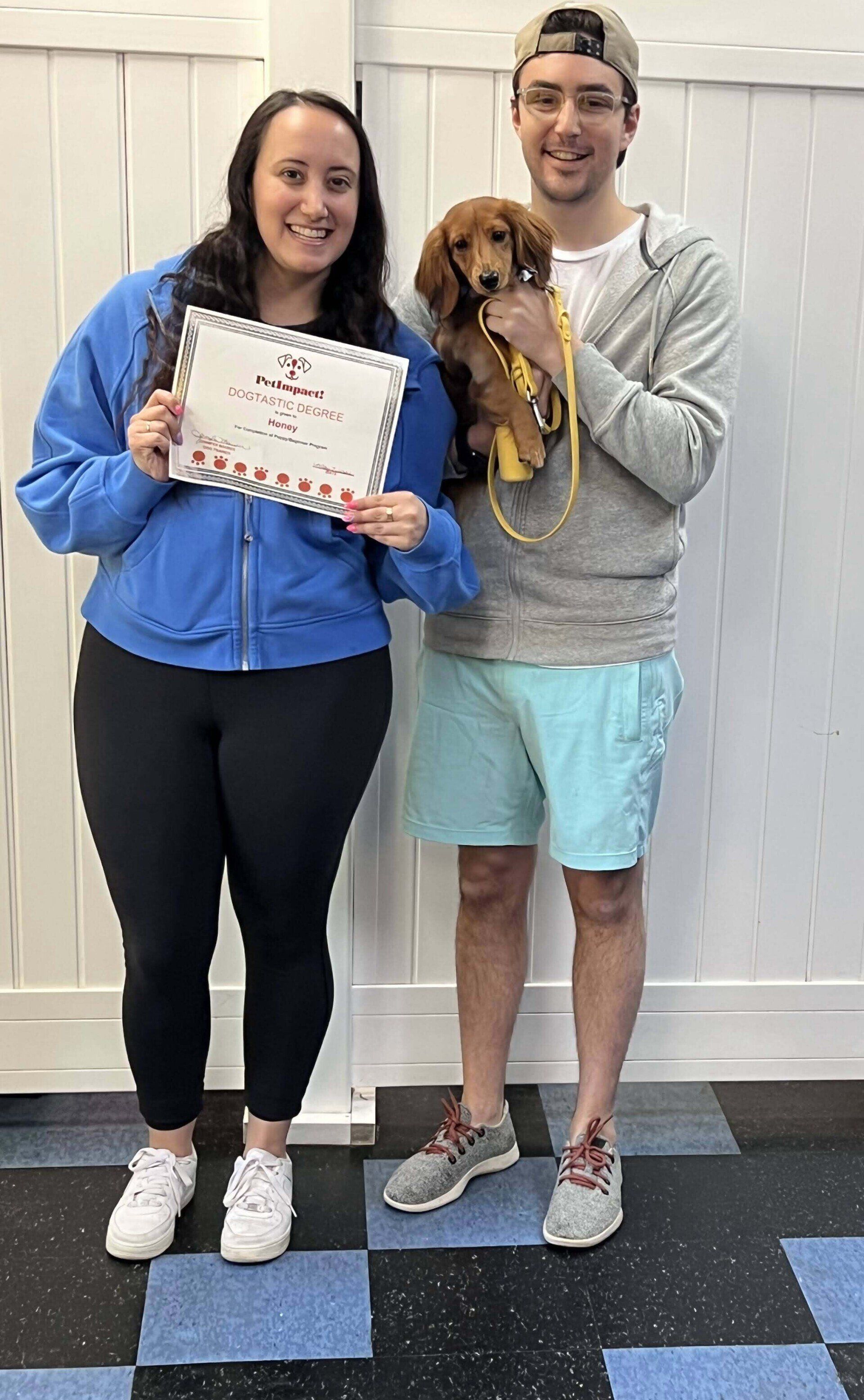 Woman and man with a dog, holding a certificate. They are smiling, indoors, with a white fence behind.