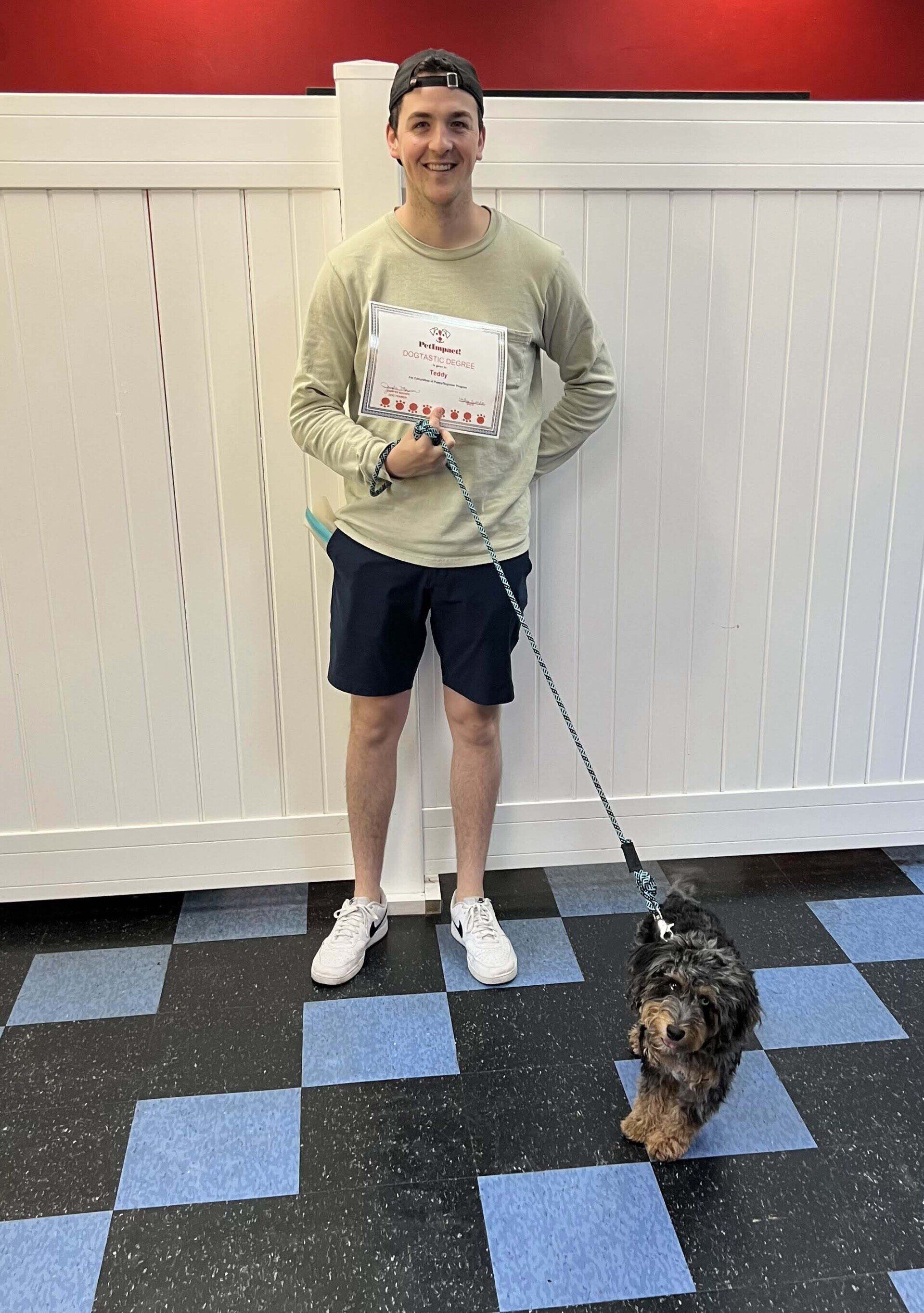 Man in shorts holding a paper, standing with a dog on a leash in front of a white fence.