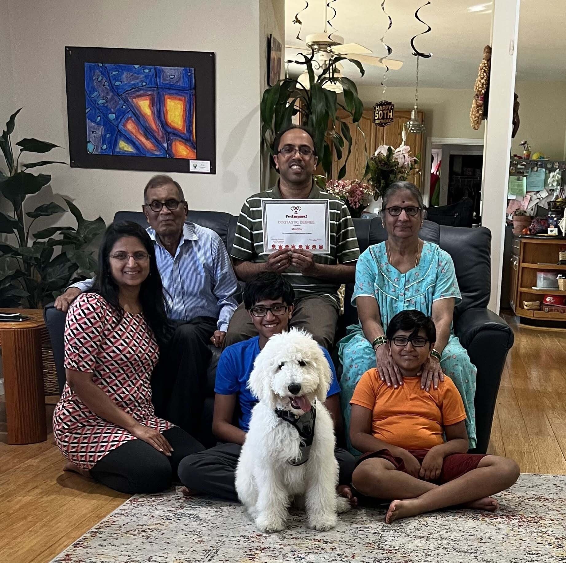 Family indoors with a fluffy white dog, smiling. A man holds a certificate. Art on the wall.