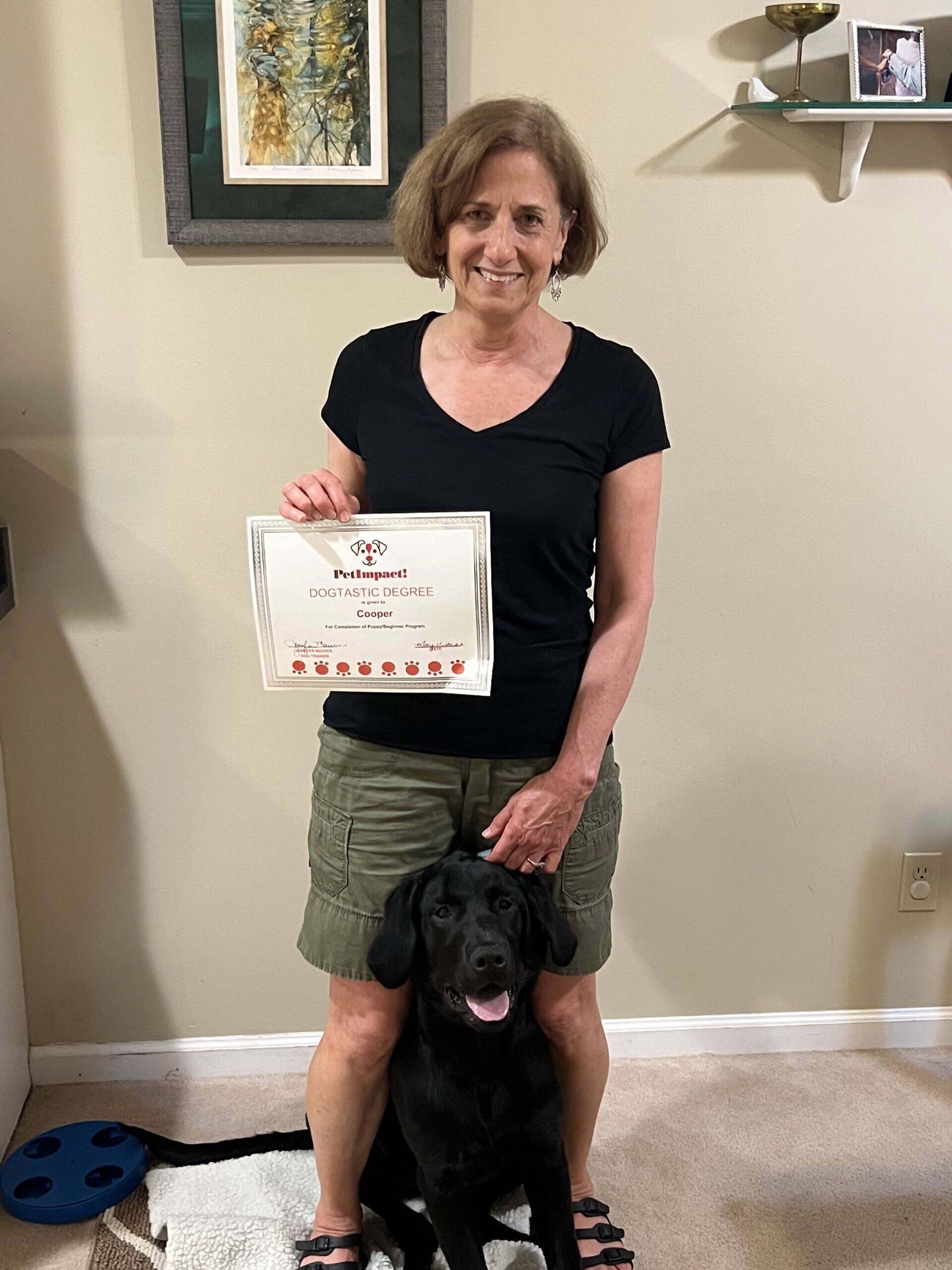 Woman in black shirt and green shorts, smiles while holding certificate, black dog sits in front.
