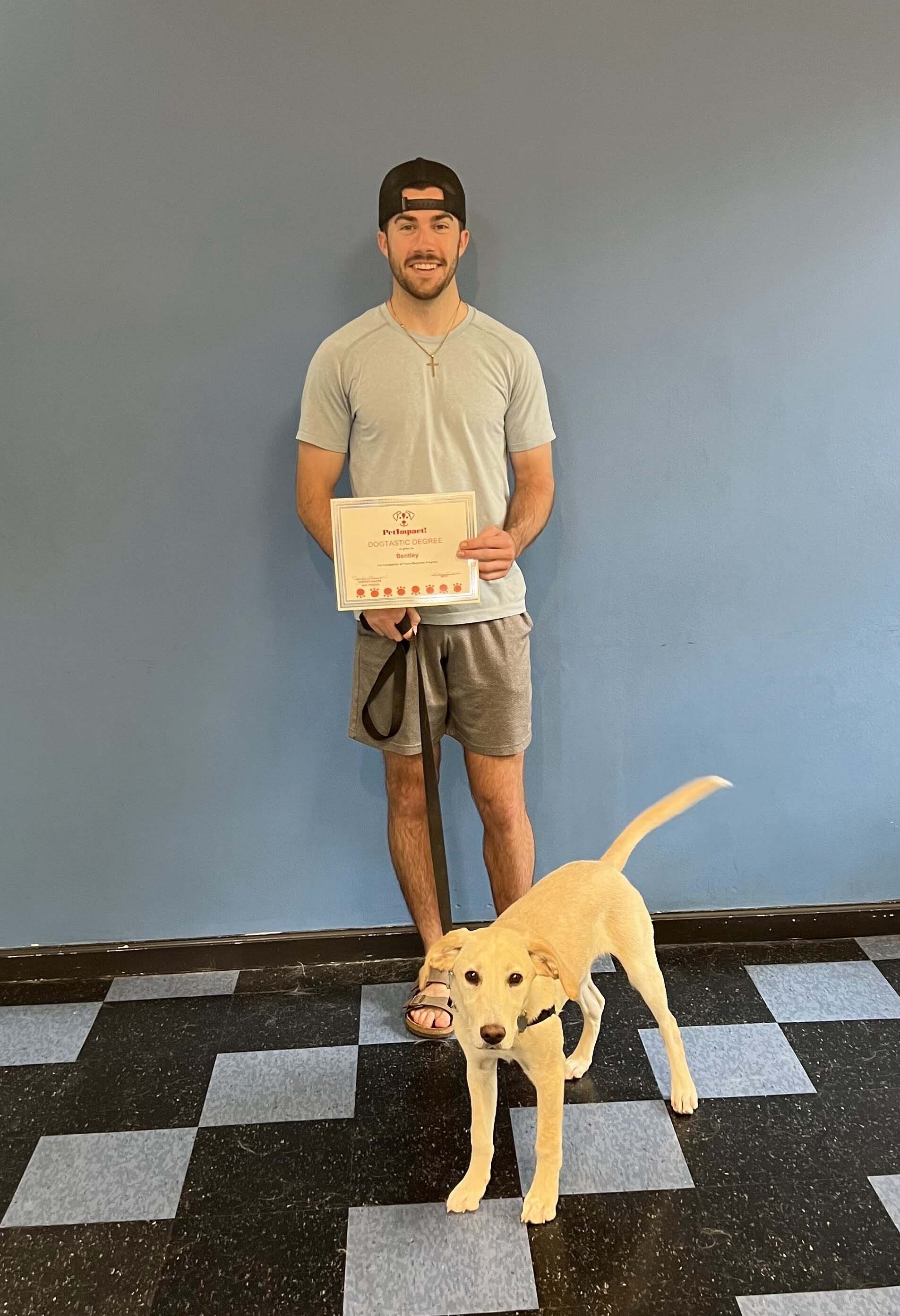 Man with certificate stands next to a yellow dog, both looking at camera. Blue wall backdrop.