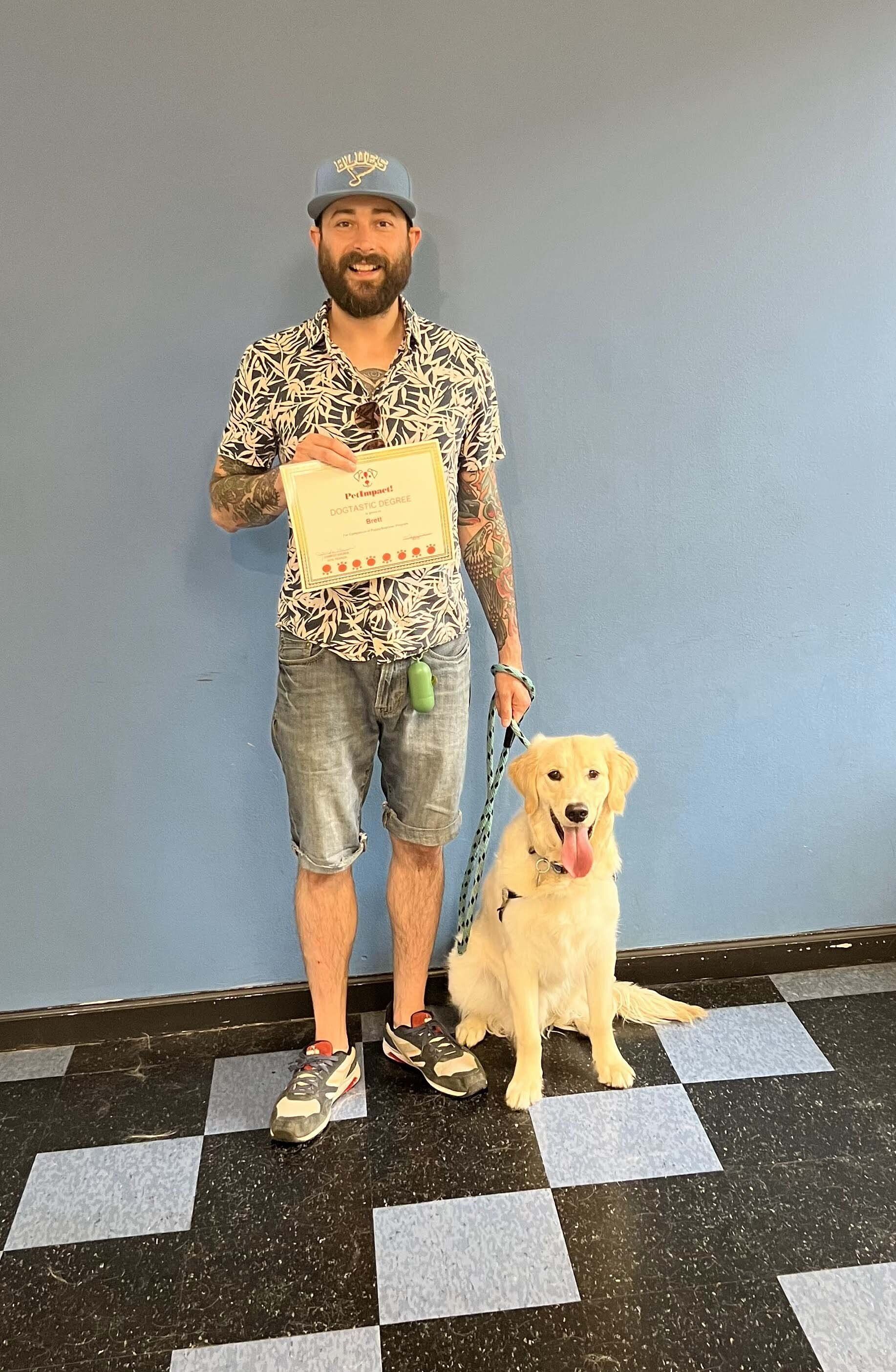 Man with tattoos and Golden Retriever dog hold certificate; posing in front of blue wall and checkered floor.