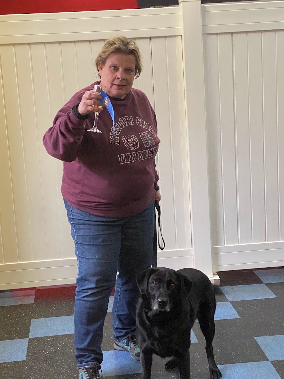 Woman with a wine glass, holding a dog's leash, standing with a black Labrador in front of a white fence.