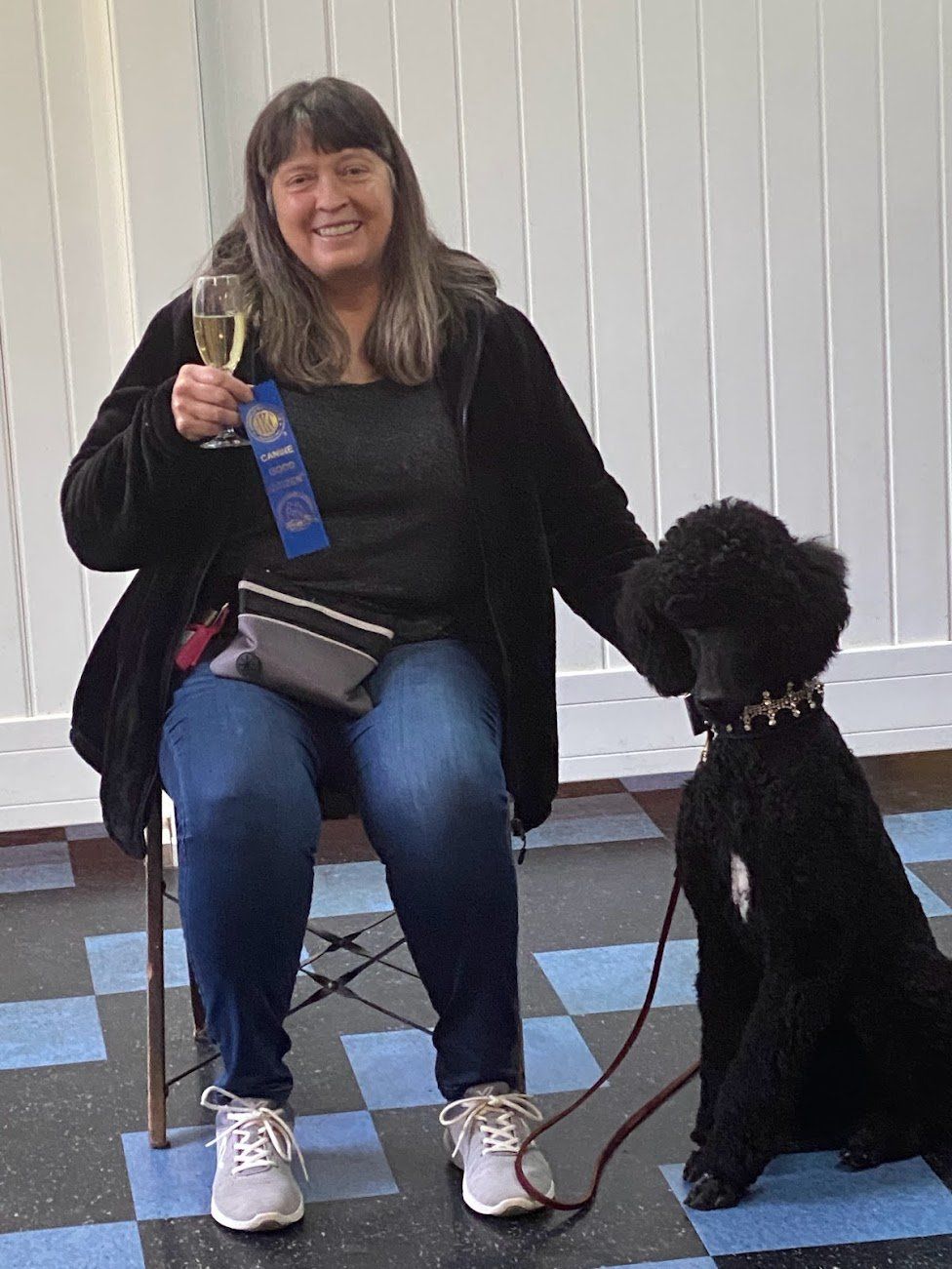 Woman and black poodle celebrate with champagne. Woman seated, holding blue ribbon.