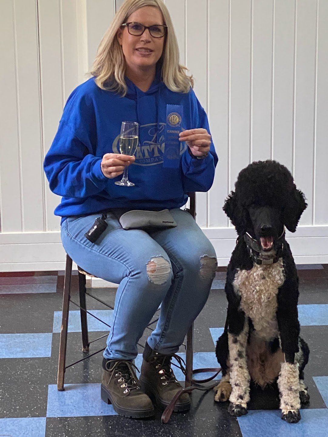 Woman in blue hoodie and jeans sits with poodle, holding a glass and a trophy.