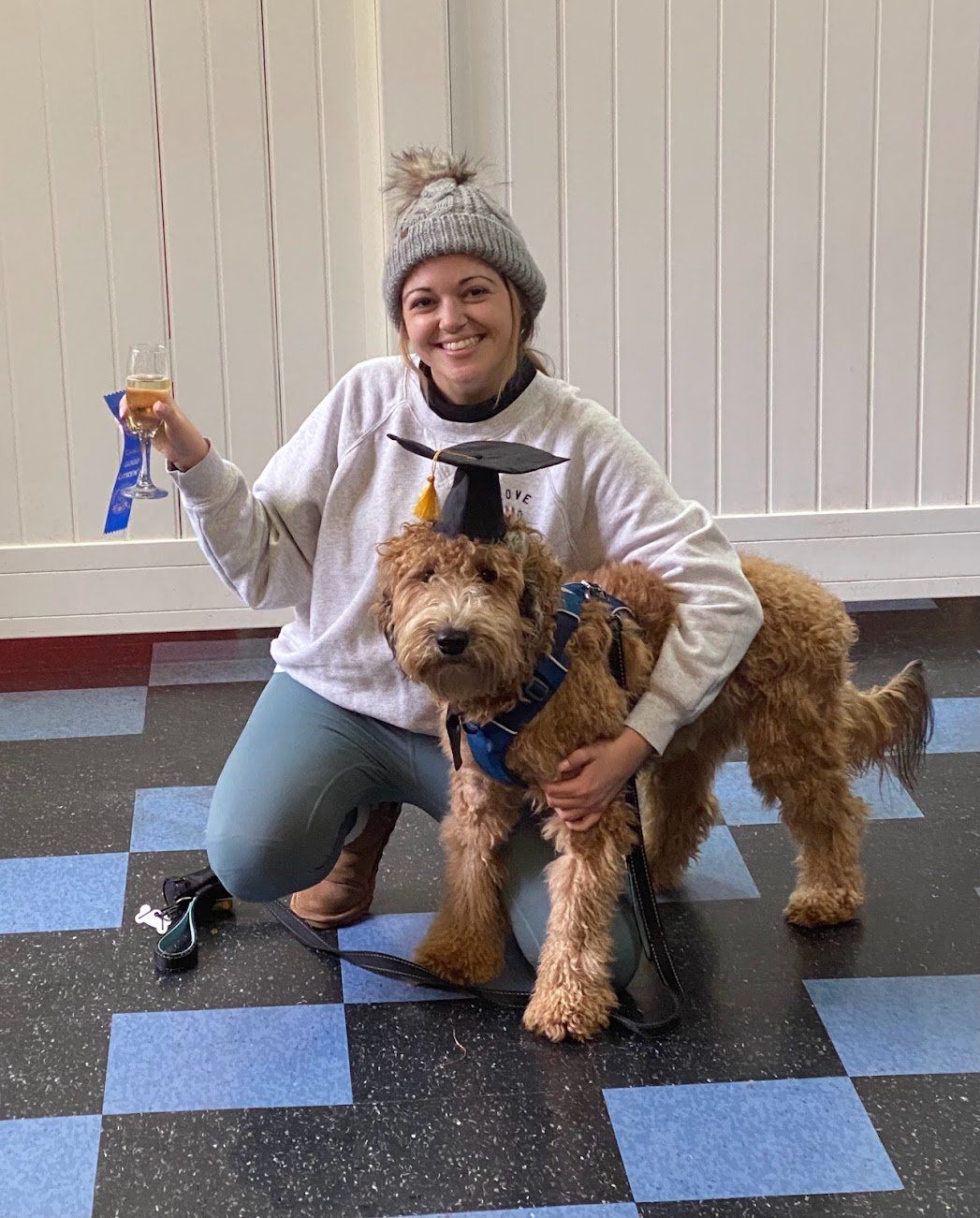 Woman kneels with a dog wearing a graduation cap, holding a champagne flute. Blue ribbon on the wall.