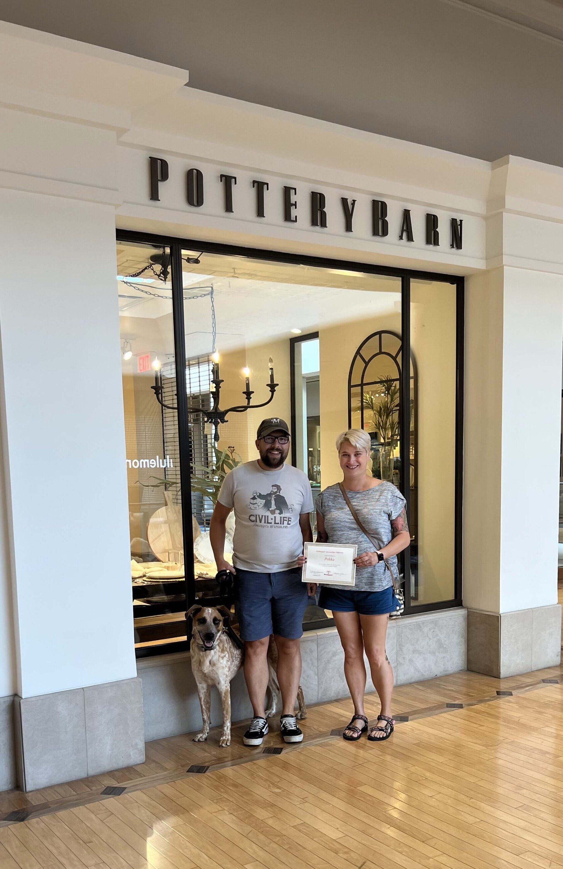 Couple and dog pose in front of a Pottery Barn store. They hold a document. Neutral setting.