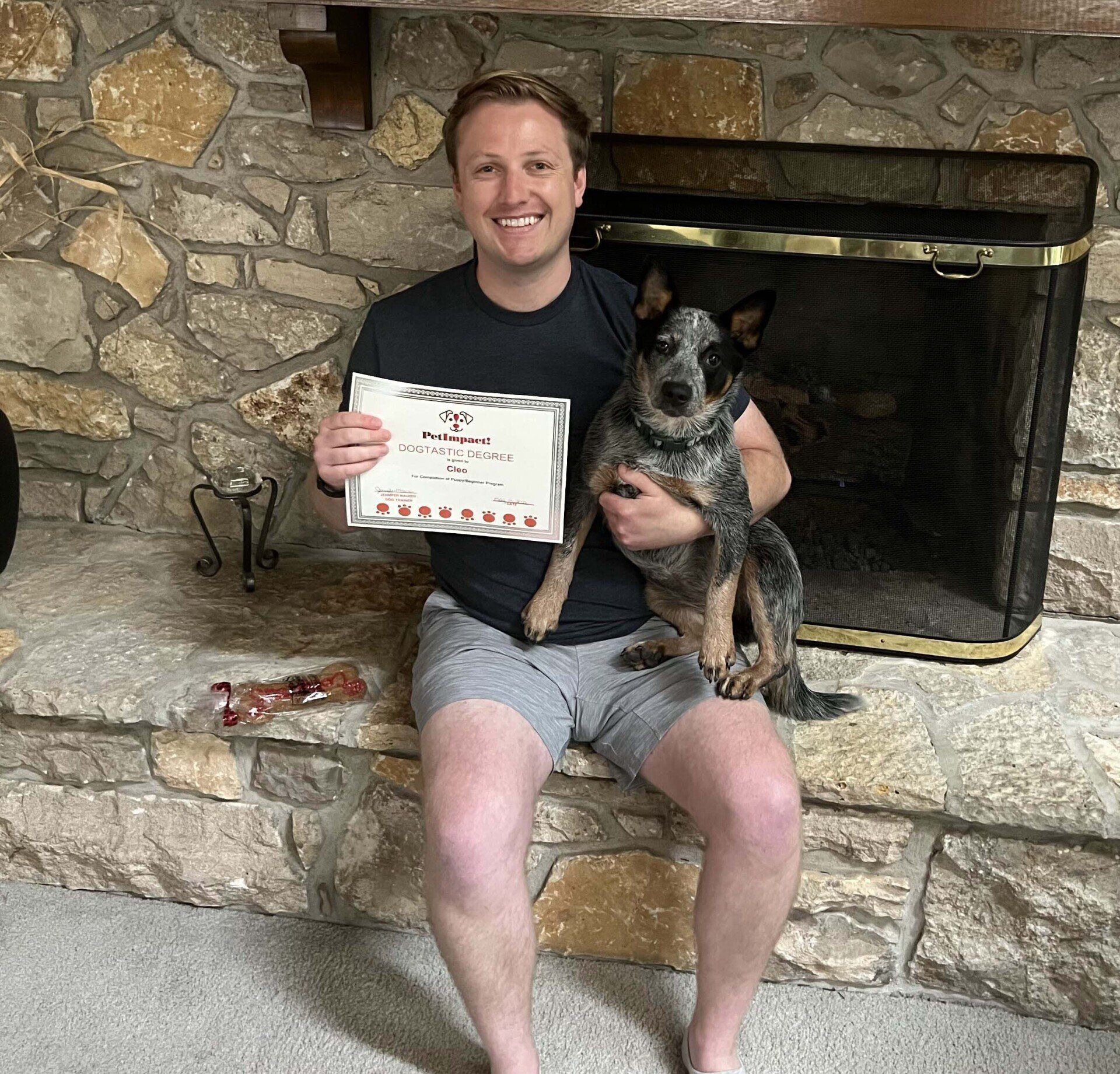 Man sitting by fireplace holding a dog and a certificate; both smile.