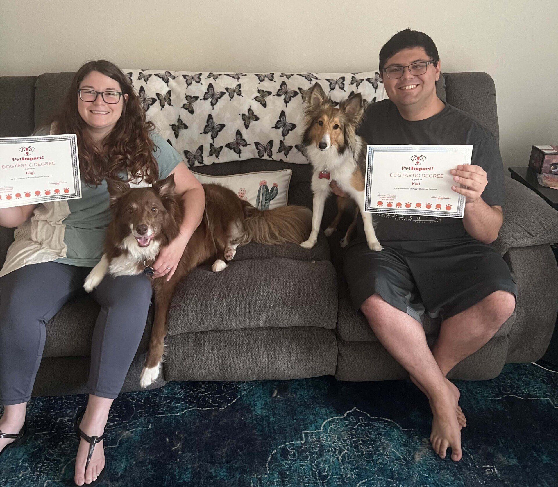 Two people and two dogs on a couch holding award certificates. The people smile.