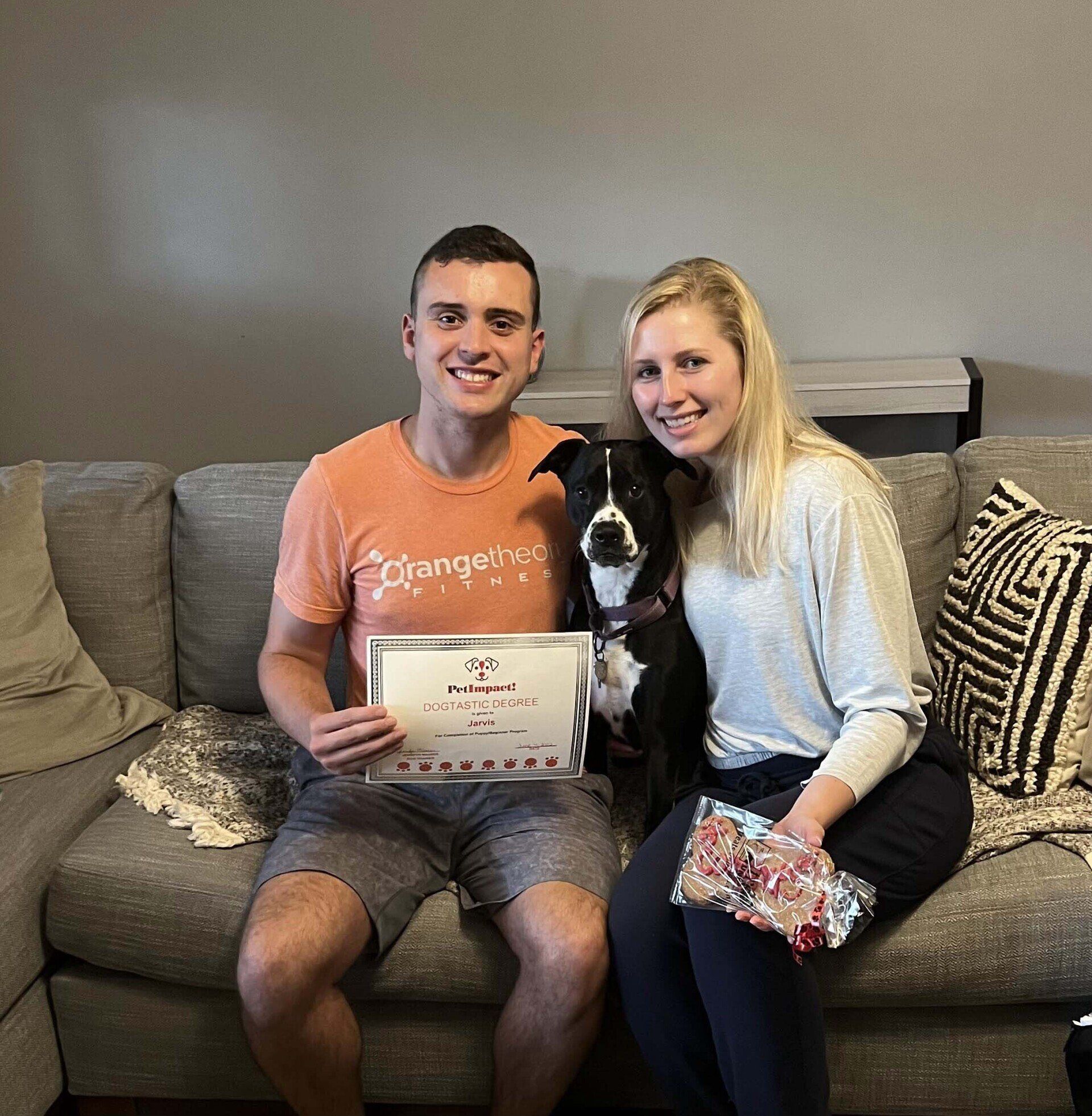 A couple and dog seated on a couch; man holds certificate. The woman holds a gift.