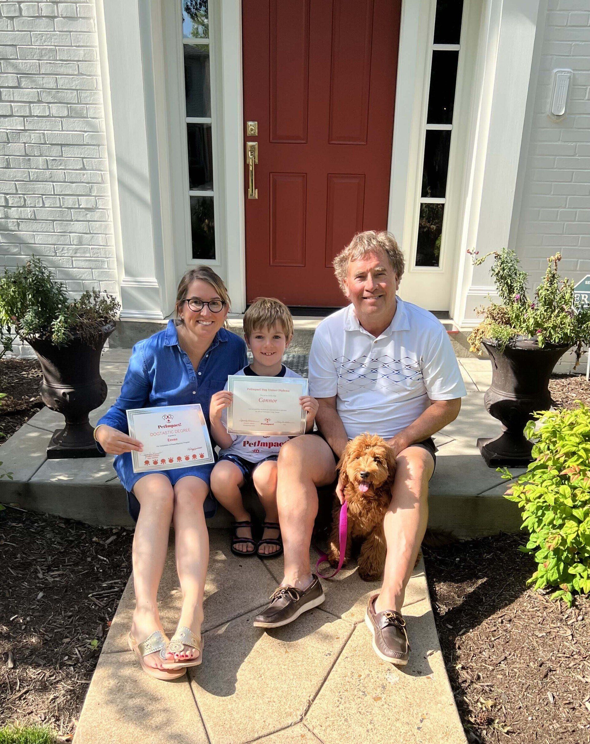 Family sitting on front steps, holding certificates. A dog sits beside the man. Red door, gray house.