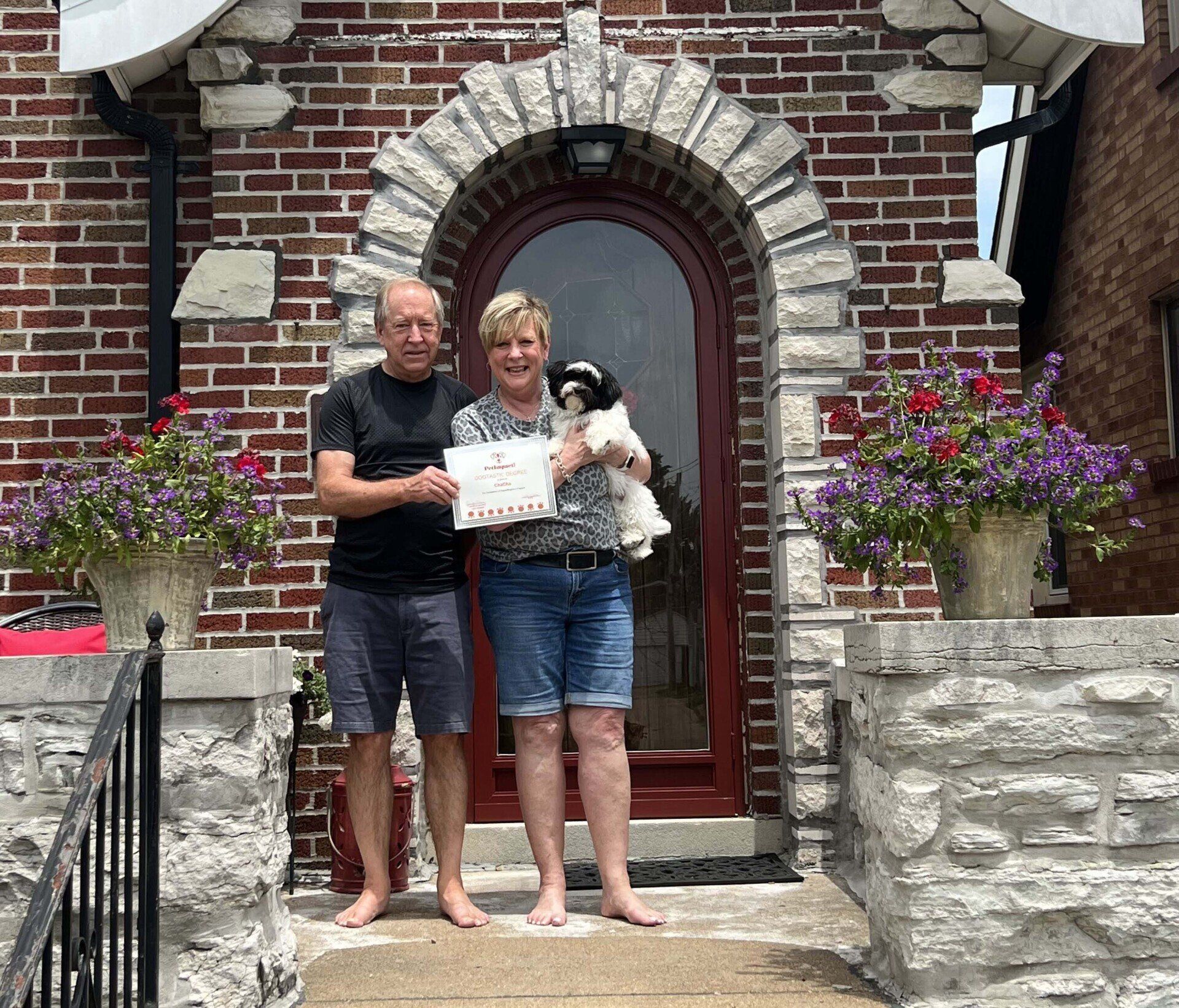 Couple and dog holding sign, standing in front of red door with stone archway, flowers in pots.