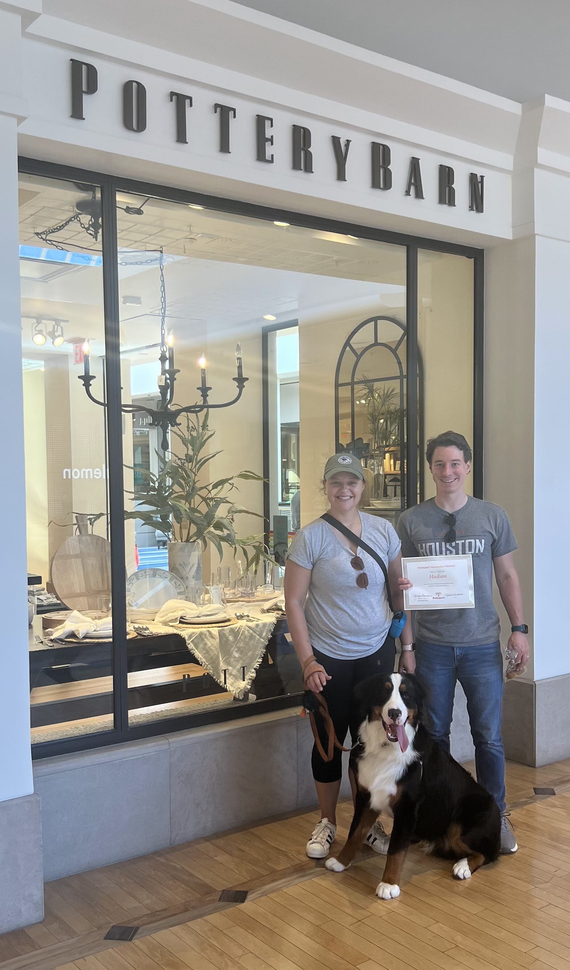 People and dog posing in front of a Pottery Barn store. They hold a white object; brick and glass facade.