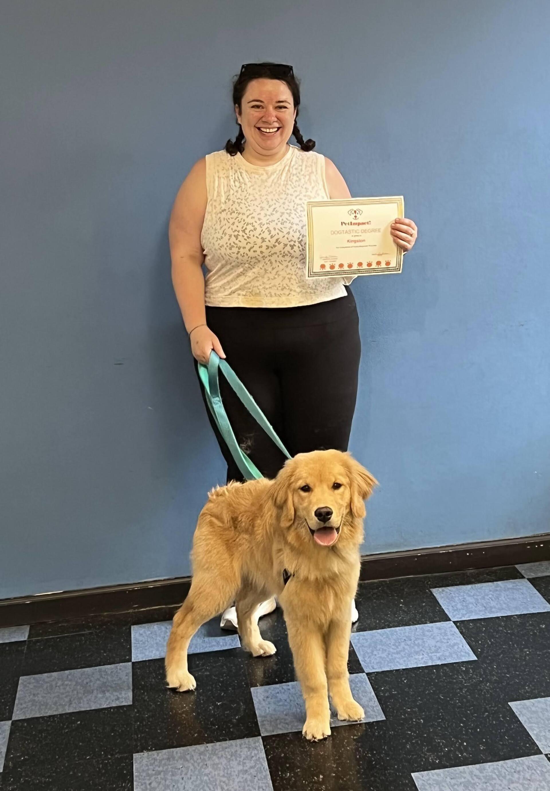 Woman smiles with Golden Retriever; holding a certificate. Blue wall and checkered floor.