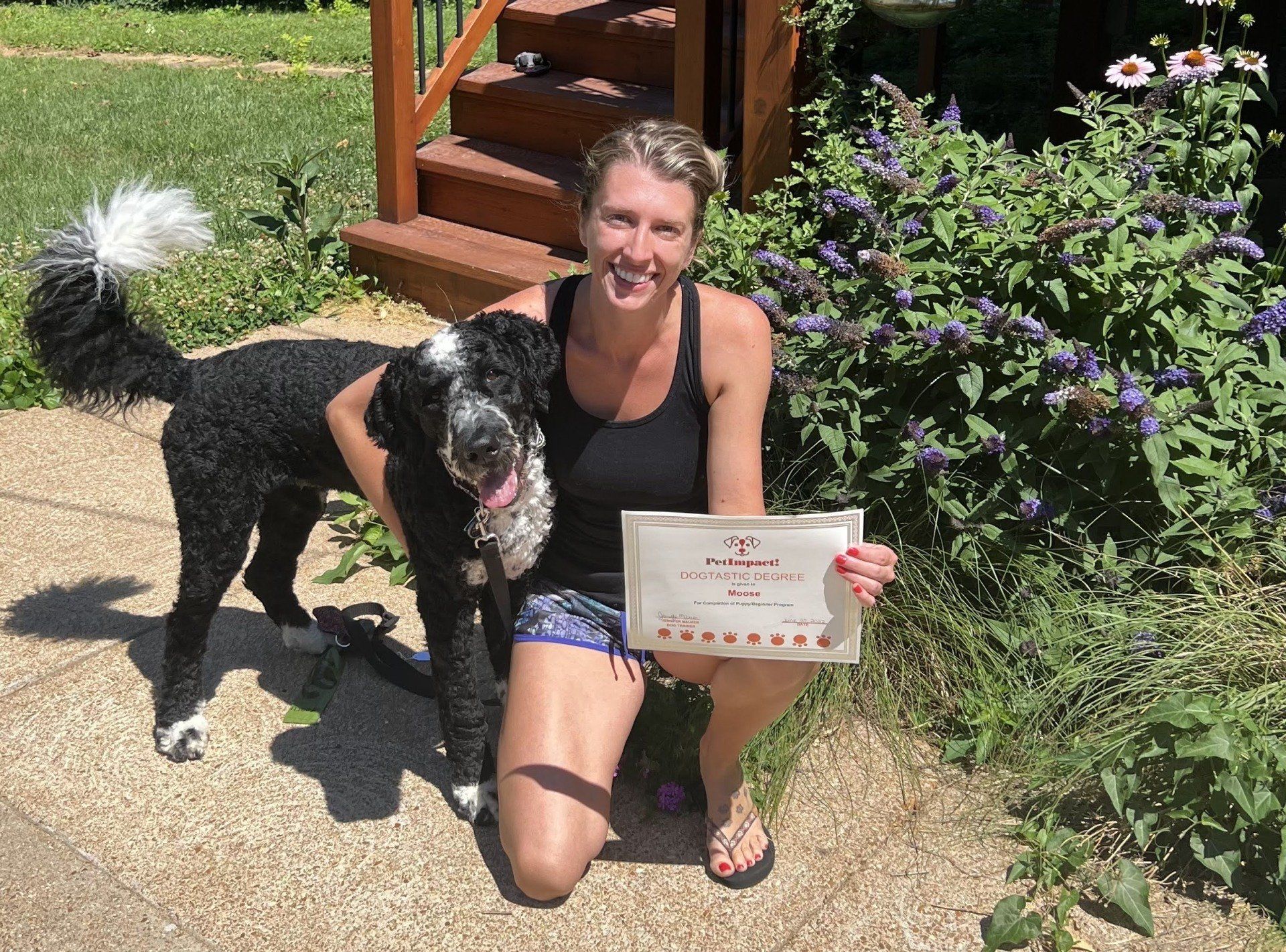 Woman kneels with a black and white dog, holding a certificate outdoors. Smiling near greenery and steps.