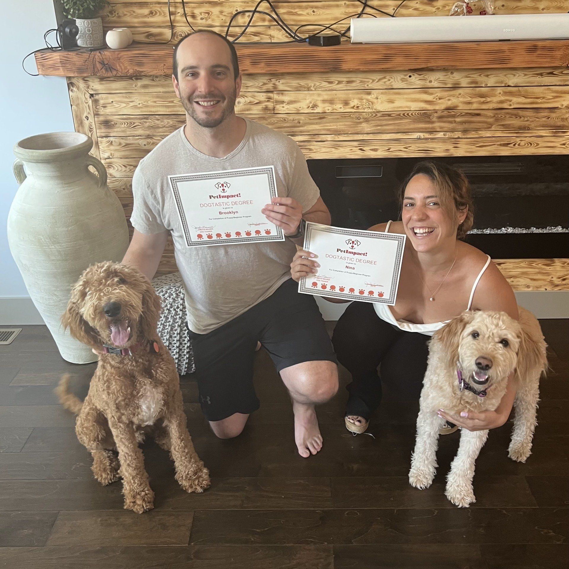 Couple kneels with two dogs, holding awards in front of a fireplace.