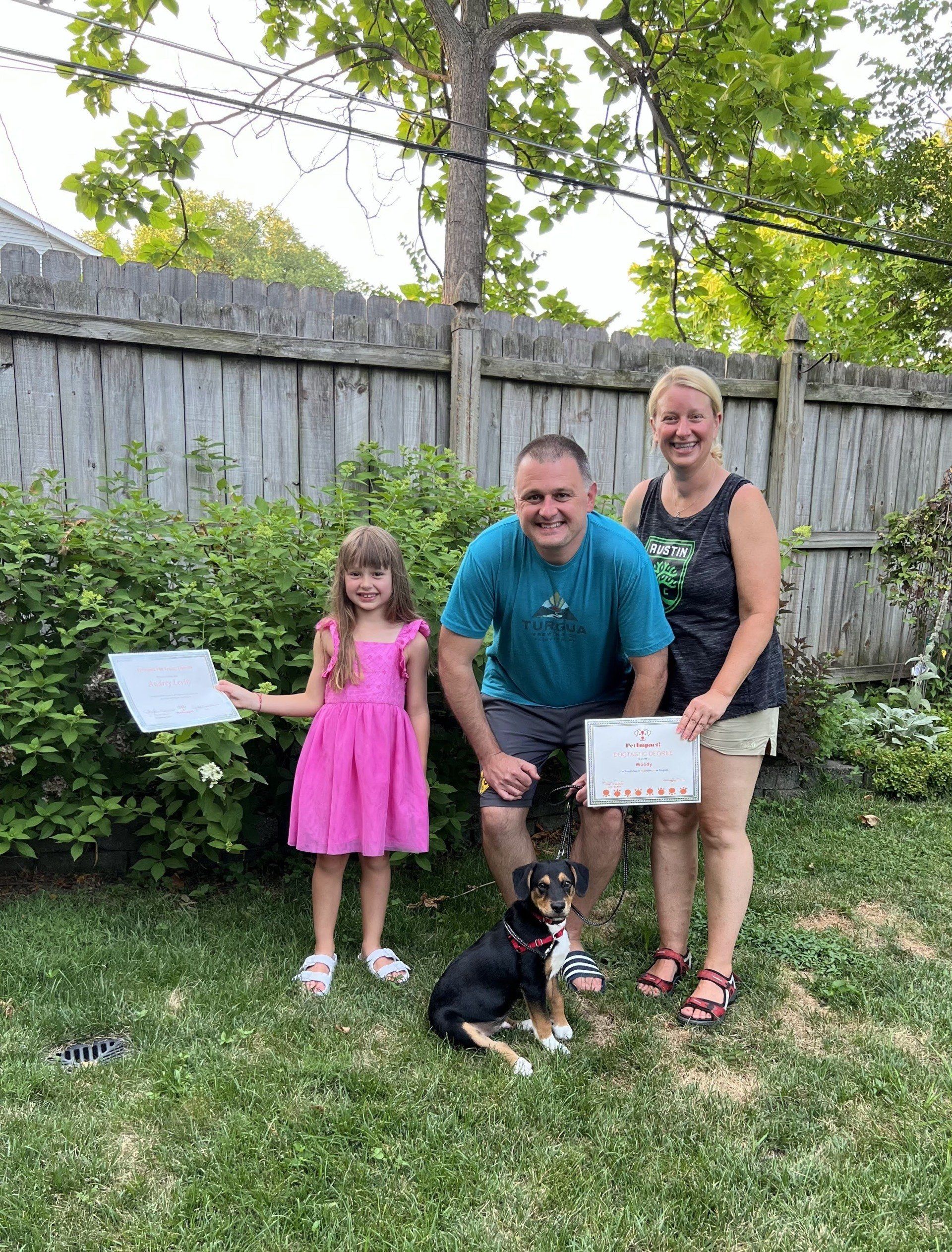 Family with a dog in a backyard, holding papers. The girl wears pink, the man turquoise, and woman black.