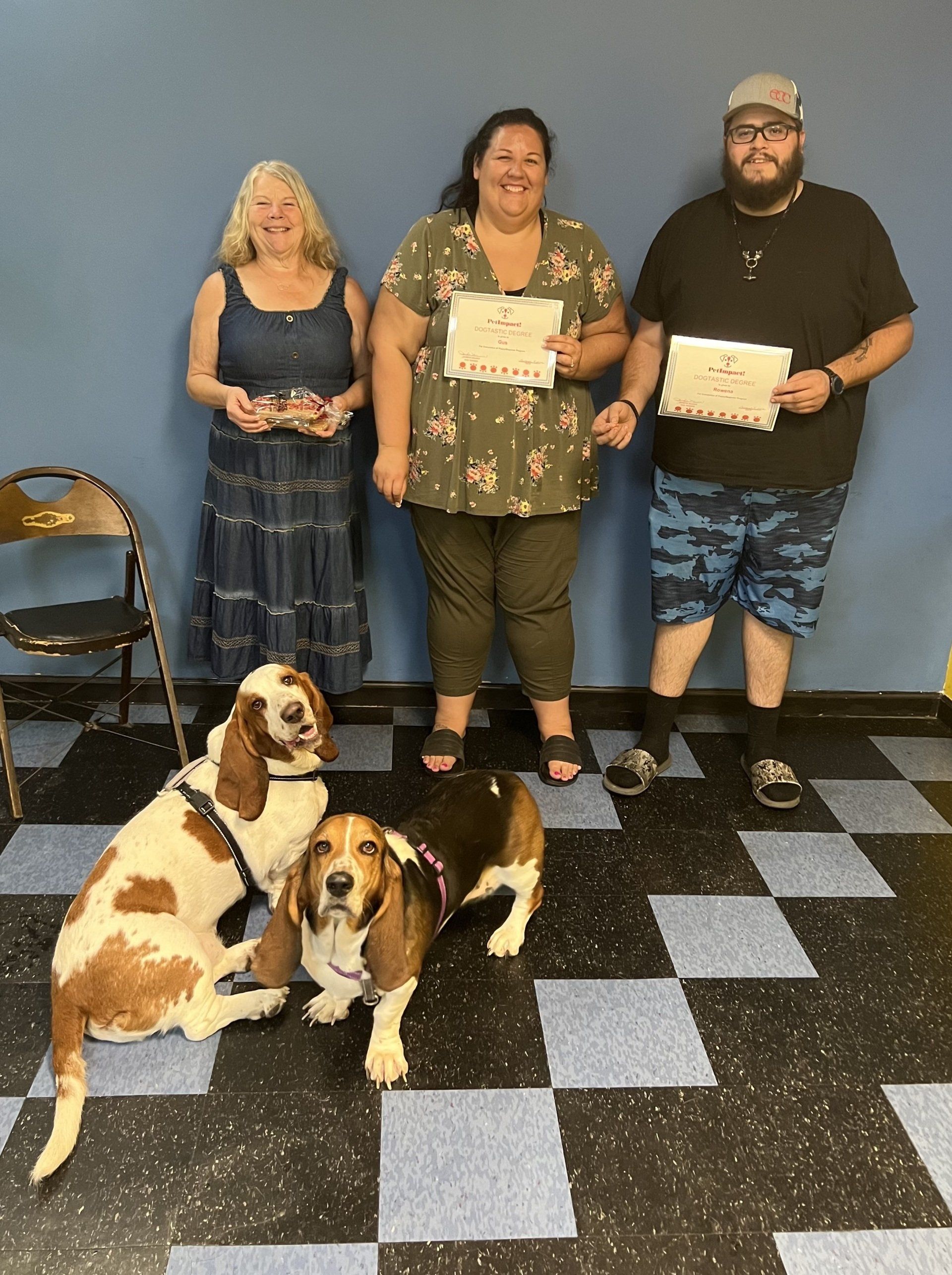 Three people and two dogs stand in front of a blue wall. The people hold certificates. Checkered floor.