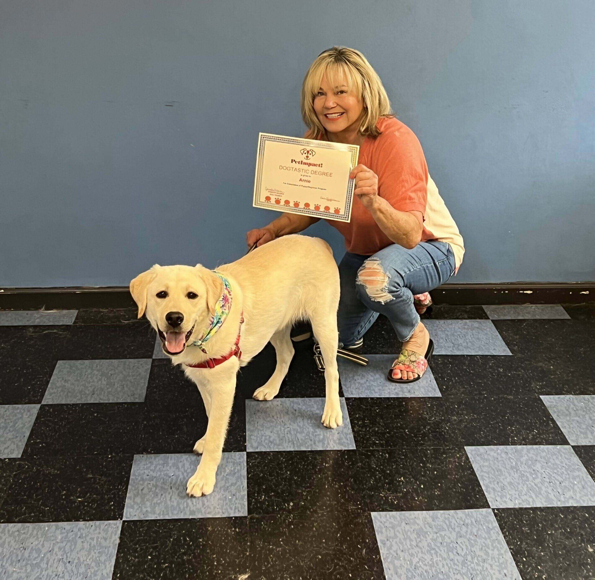 Woman with blonde hair kneels beside a yellow Labrador dog holding a certificate; both smile.