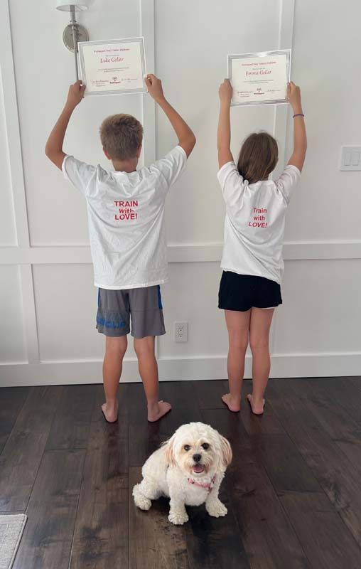 Two children holding certificates, standing near a small dog, wearing matching shirts, in front of a white wall.