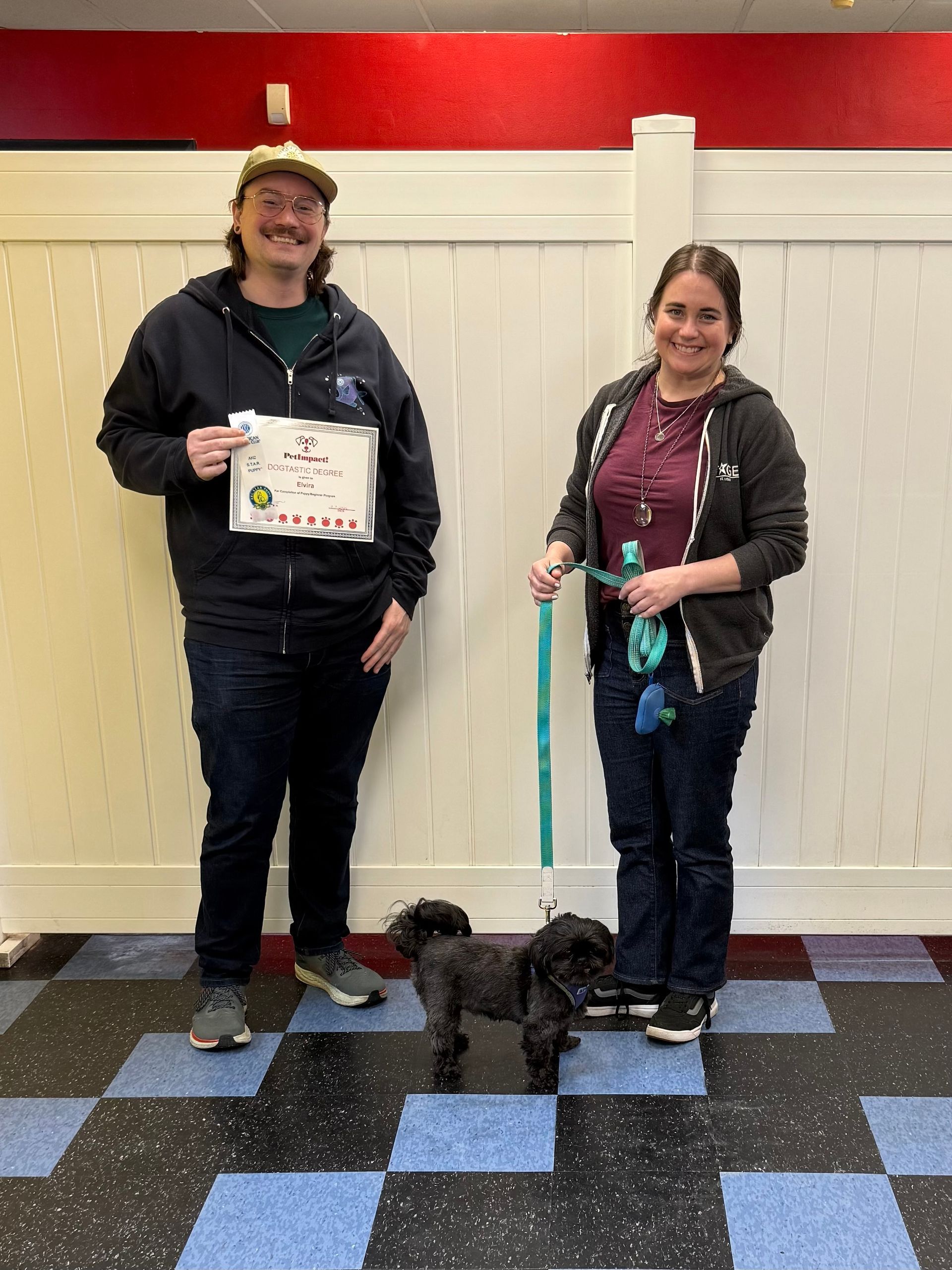 Two people stand with a small black dog on a leash in front of a white fence; one holds a certificate.