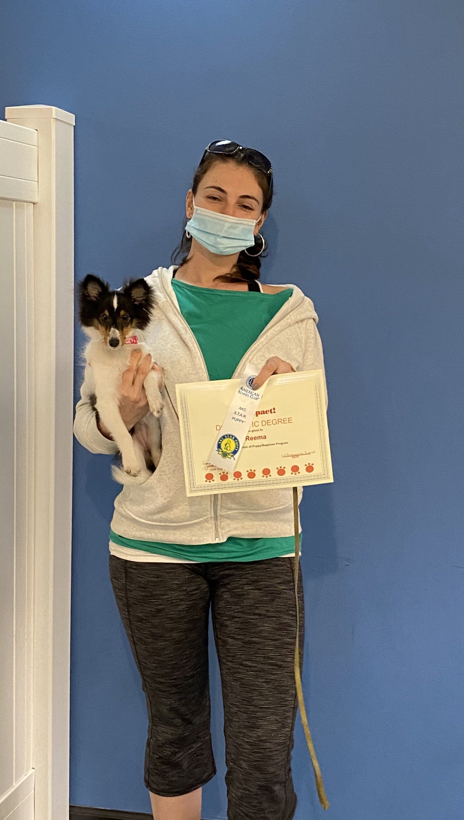 Woman in mask holds dog and certificate; blue wall.