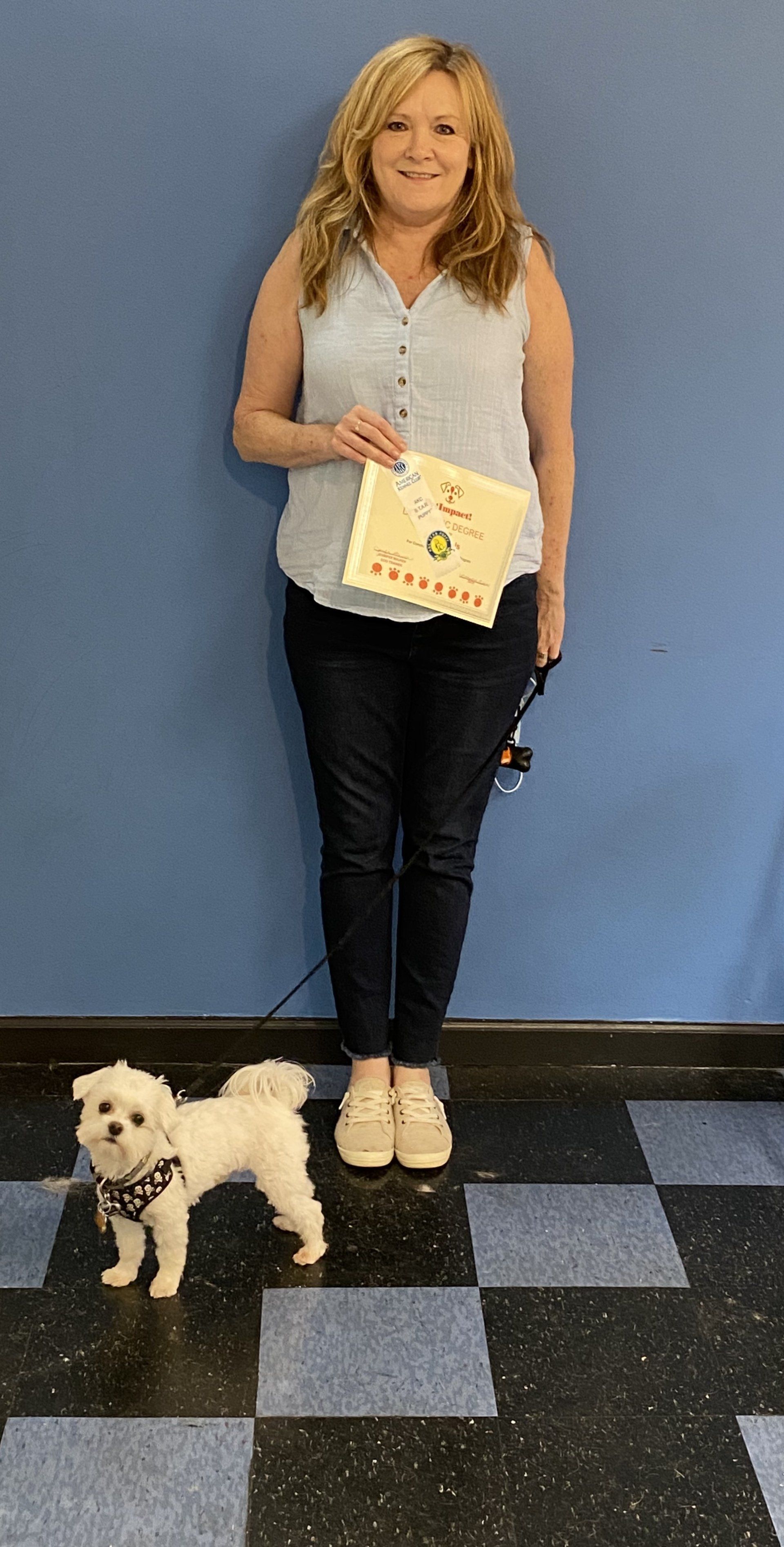 Woman holding a certificate, standing with a small white dog; against blue wall, checkered floor.