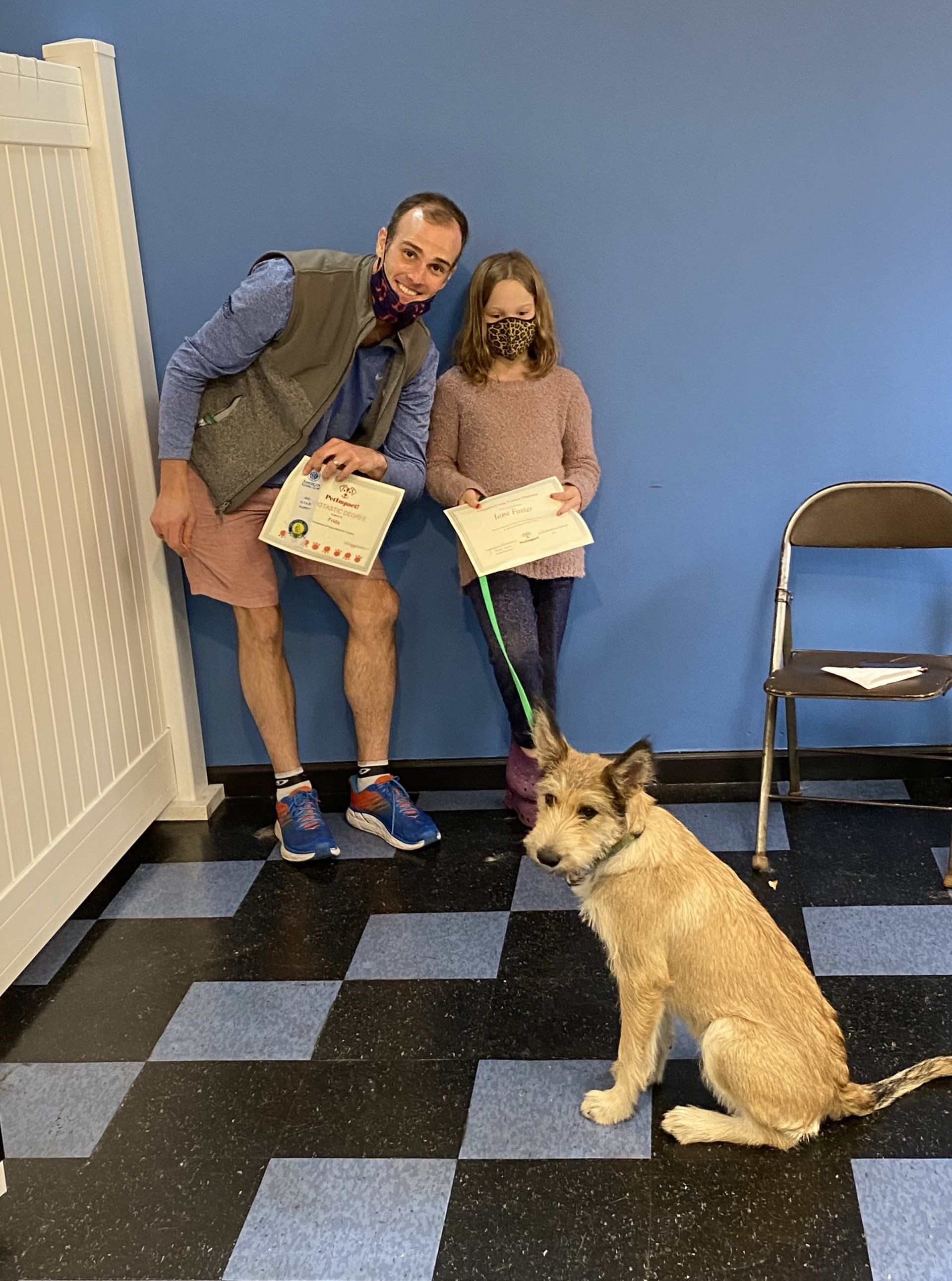 Man, girl, and dog pose, holding certificates near a blue wall and checkered floor.