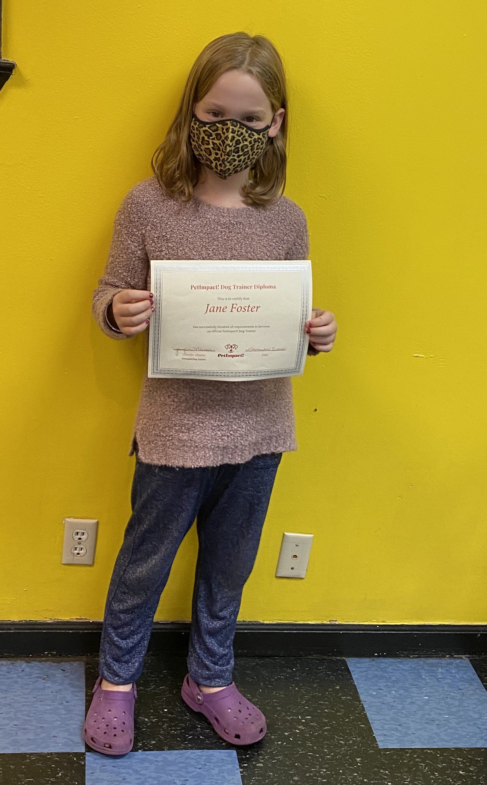 Girl in mask holding a certificate, standing against a yellow wall, wearing purple shoes and jeans.