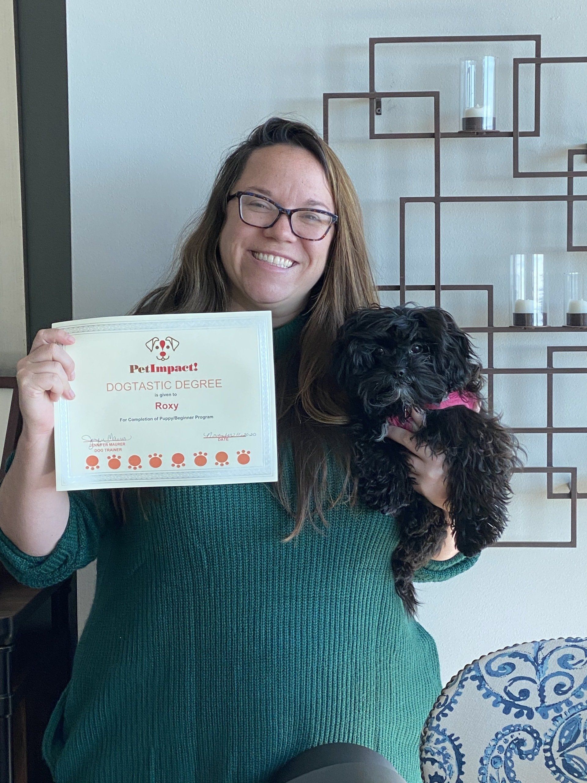 Woman in green sweater holding a certificate and black dog with pink collar, smiling. Decorative wall in background.